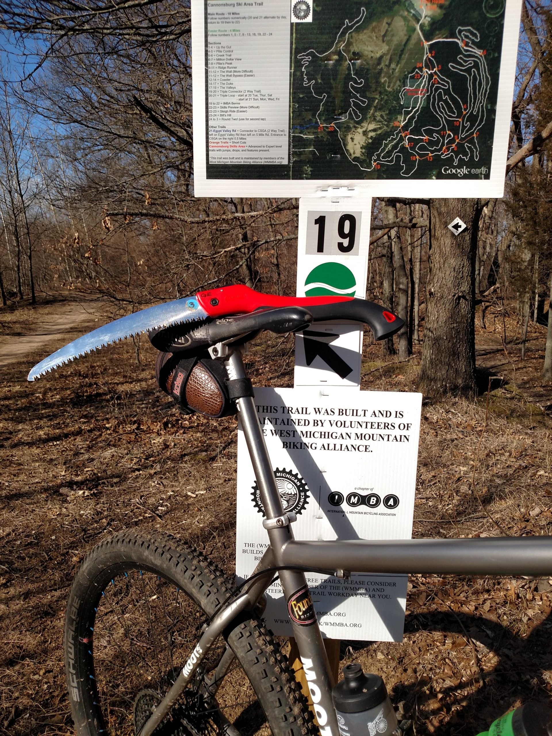 A mountain bike parked beside a trail sign that provides information about the Cannonsburg Ski Area Trail. The sign includes a map of the trail system marked with various numbered trails and indicates it's maintained by local volunteers. A red-handled saw is visible on the bike's seat, and the surrounding area is wooded with bare trees and a clear blue sky. Cannonsburg Ski Area mountain bike trail.