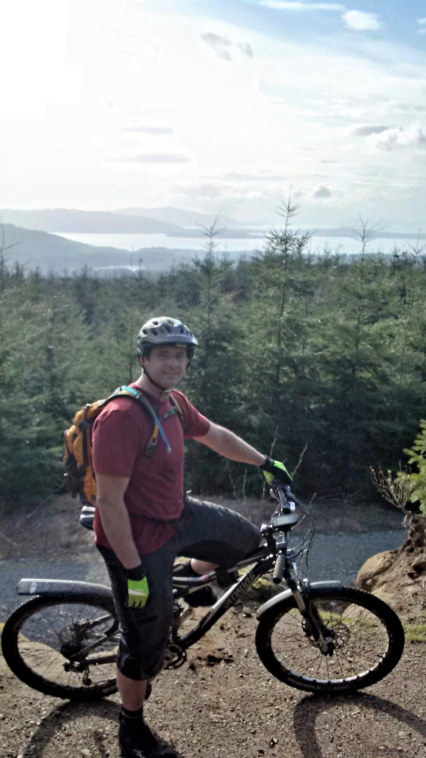 A person in a helmet poses next to a mountain bike on a rocky trail surrounded by young evergreen trees. The background features a scenic view of hills and a body of water under a partly cloudy sky, suggesting an outdoor adventure in a natural setting. Galbraith Mountain mountain bike trail.