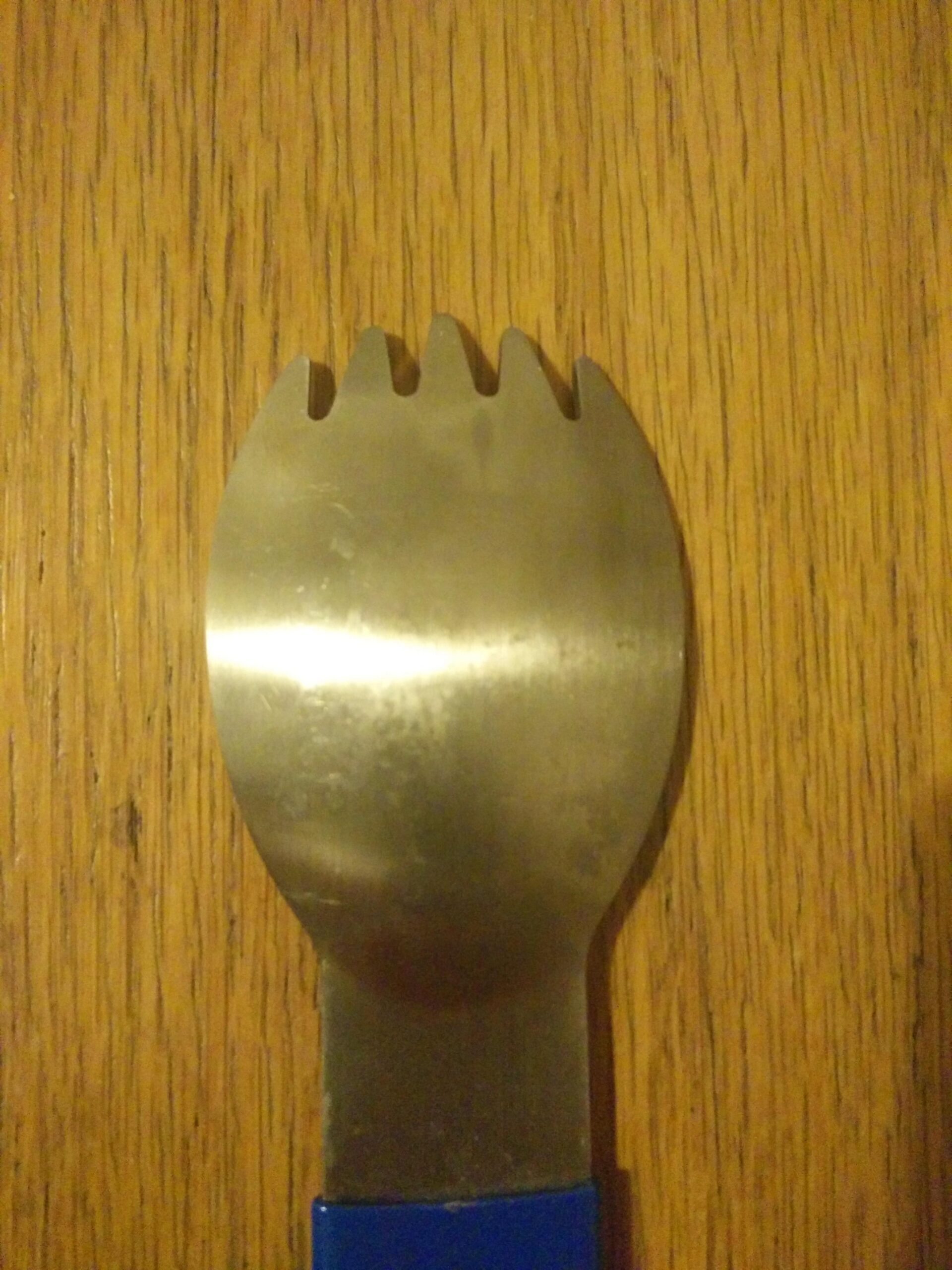 Park Tool SPK-1 Spork: A close-up image of a stainless steel spork, featuring three fork tines and a rounded scoop shape. The handle is blue plastic and the spork is placed on a wooden surface.
