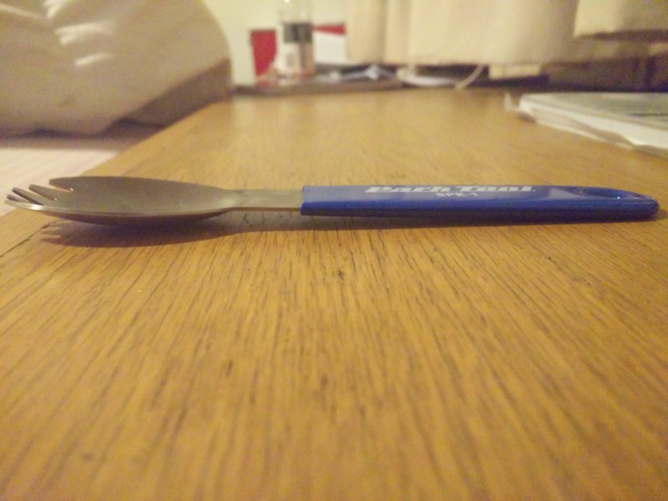 Park Tool SPK-1 Spork: A close-up view of a utensil, specifically a fork with a blue handle, resting on a wooden table surface. The utensil is designed for compact use, showcasing a forked end and a metal prong. In the background, a blurred setting includes a part of a bed and some items on the table.