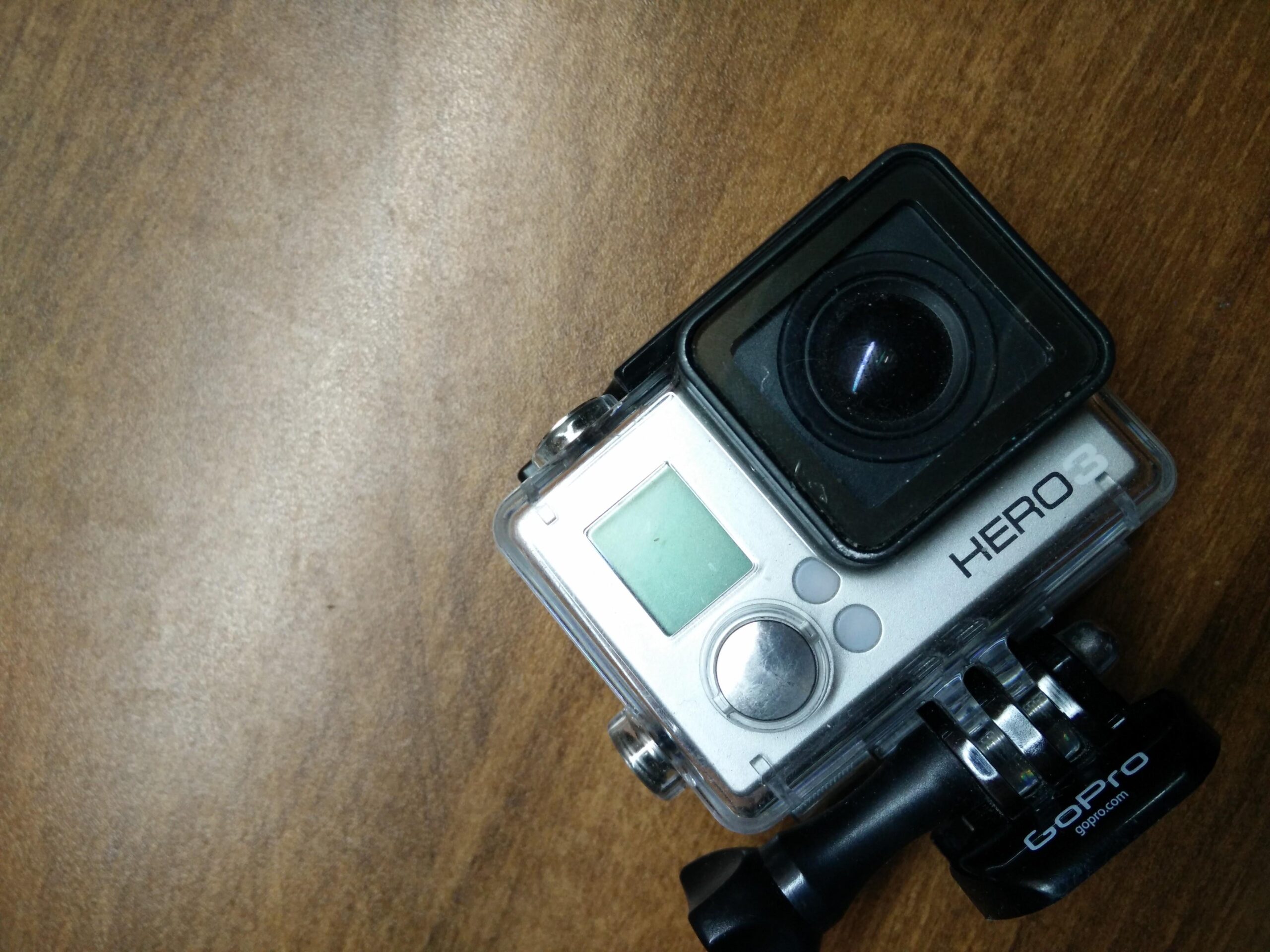 GoPro Hero 3 White Edition: A silver GoPro HERO 3 camera, partially enclosed in its protective housing, placed on a wooden table. The camera features a lens on the front, a small display screen, and control buttons on the side. A mounting bracket is visible at the bottom.