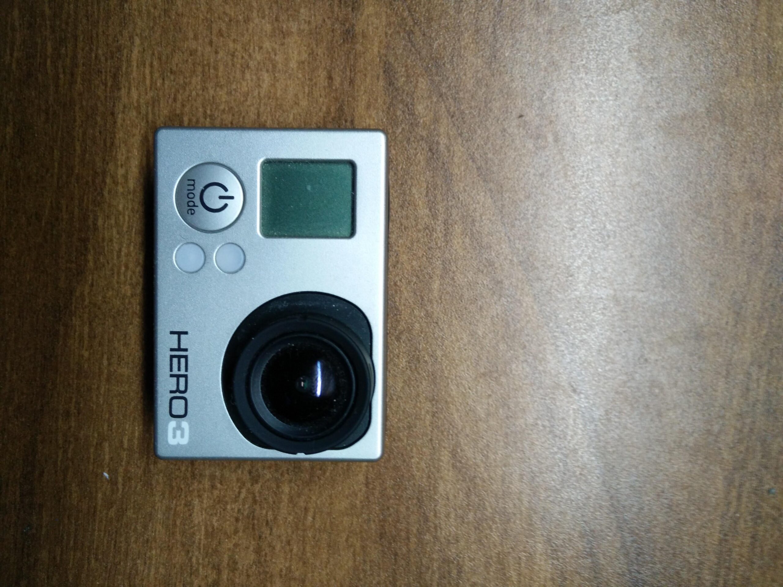 GoPro Hero 3 White Edition: A silver action camera labeled "HERO3" resting on a wooden surface. The camera features a lens, a small display screen, and several buttons, including a power button and a mode selection button.