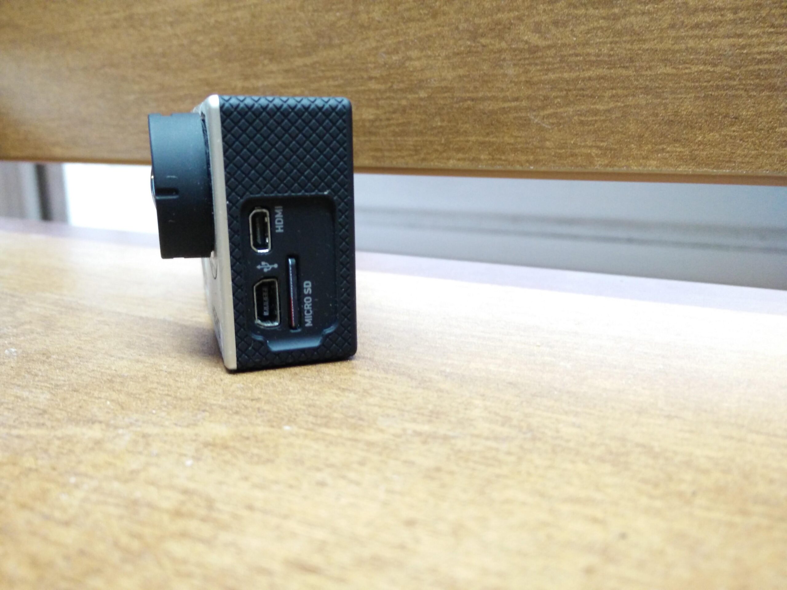 GoPro Hero 3 White Edition: A close-up view of a camera or video recording device placed on a wooden surface. The image shows the back side of the device, featuring ports labeled HDMI, USB, and Micro SD card slot.