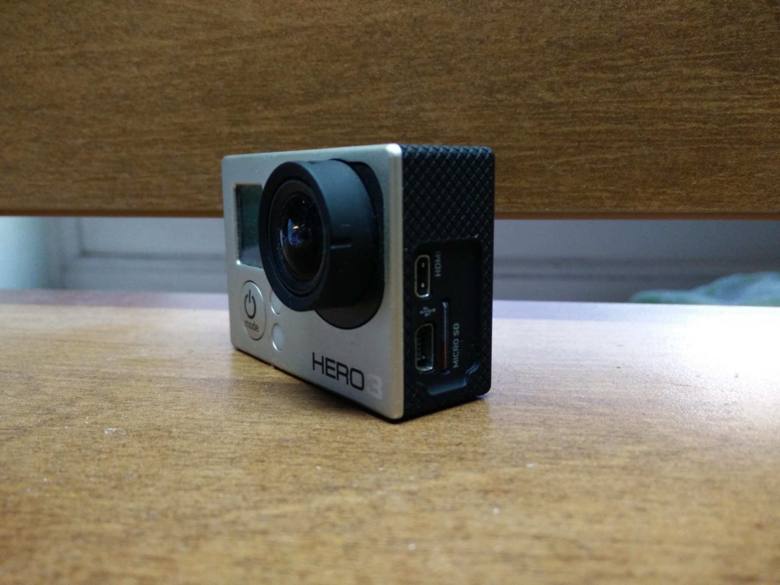 GoPro Hero 3 White Edition: A compact action camera positioned on a wooden surface, featuring a lens on the front, a power button, and connection ports for HDMI and USB. The model name "HERO3" is visible on the side of the camera.