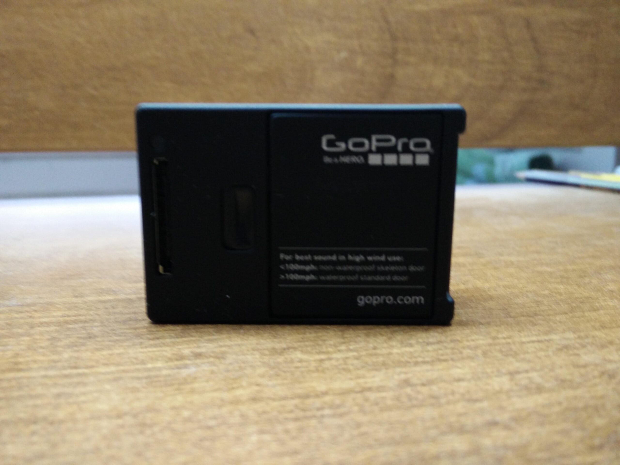 GoPro Hero 3 White Edition: GoPro camera back view, showing the card slot and labeled features. The device is placed on a wooden surface, with a textured background.