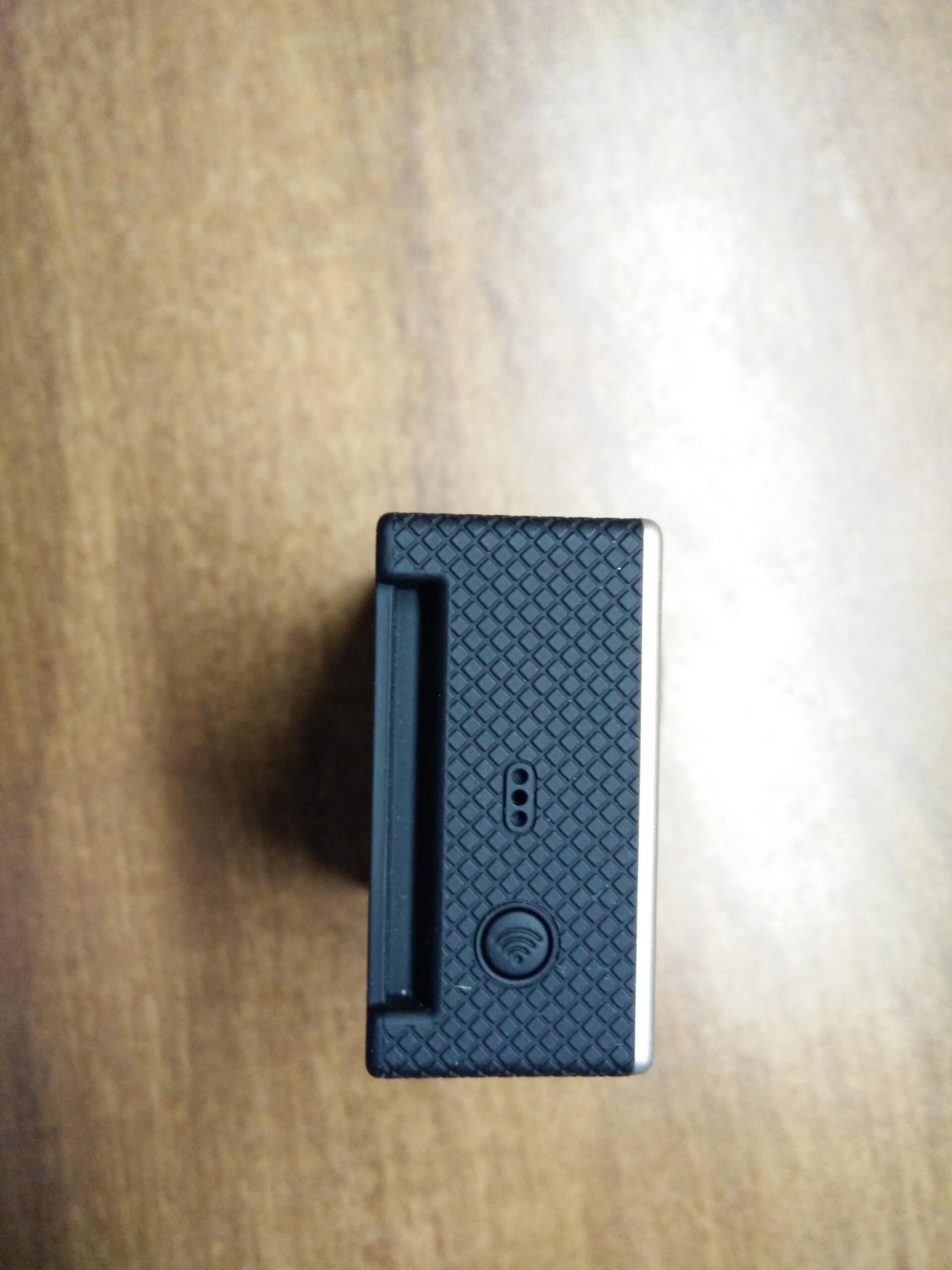 GoPro Hero 3 White Edition: Top view of a small black electronic device with a textured surface, featuring a slot and a wireless connectivity symbol on the side. The device is resting on a wooden surface.