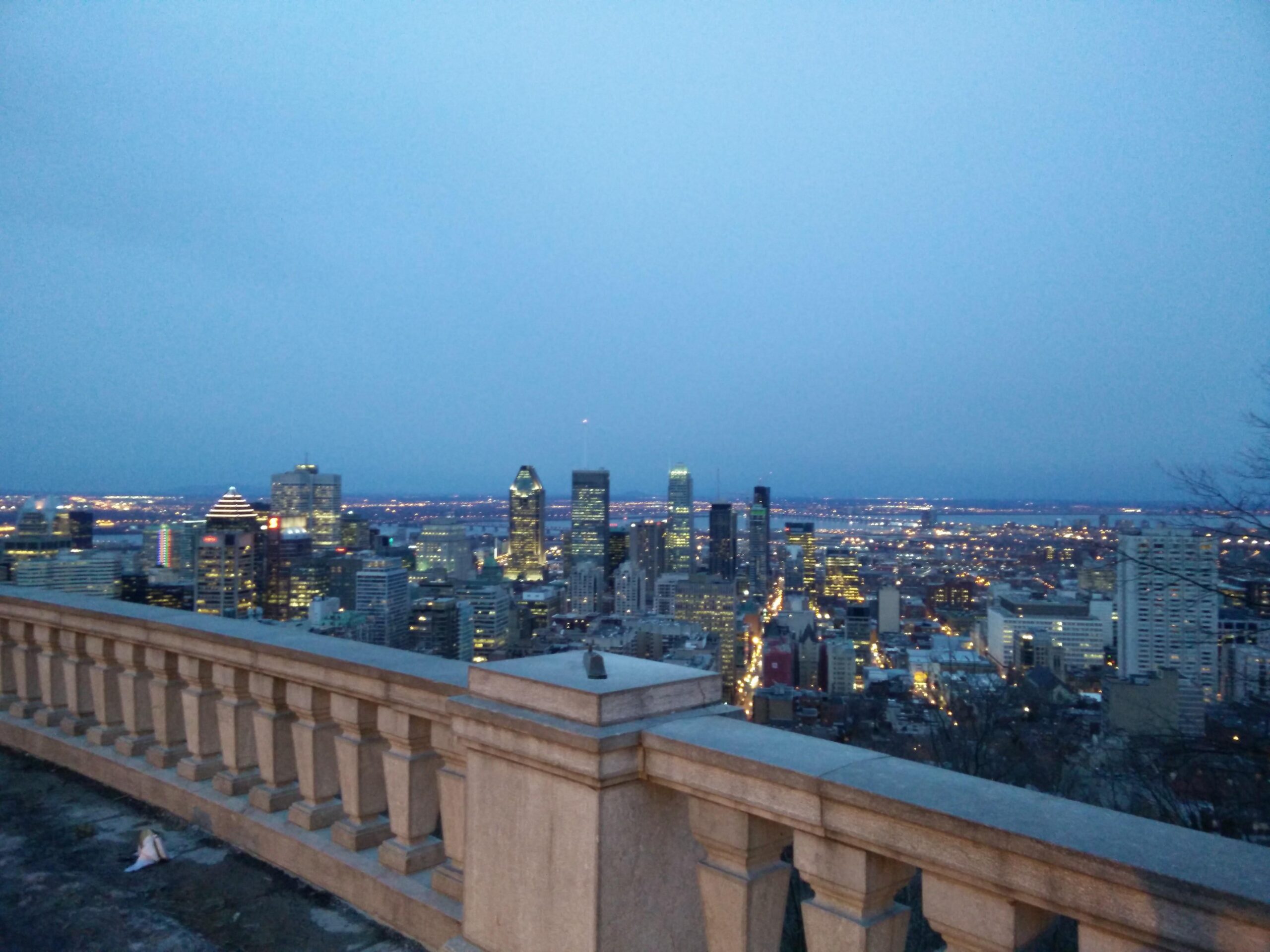 A view of downtown Montreal at dusk, with the skyline illuminated and a soft blue sky above. A stone railing in the foreground frames the city, showcasing various buildings and bright lights reflecting the evening ambiance. Parc du Mont-Royal mountain bike trail.