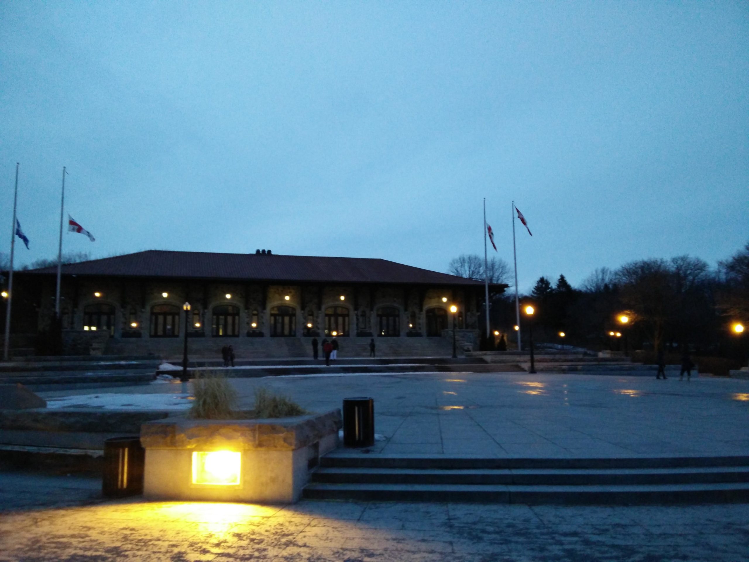 A dimly lit stone building with a red-tiled roof, flanked by flags, is set against a twilight sky. The entrance features multiple windows, and the surrounding area includes lampposts illuminating the scene. Several people are visible walking in the plaza, which has a smooth, reflective surface, suggesting recent rain or melting snow. Parc du Mont-Royal mountain bike trail.