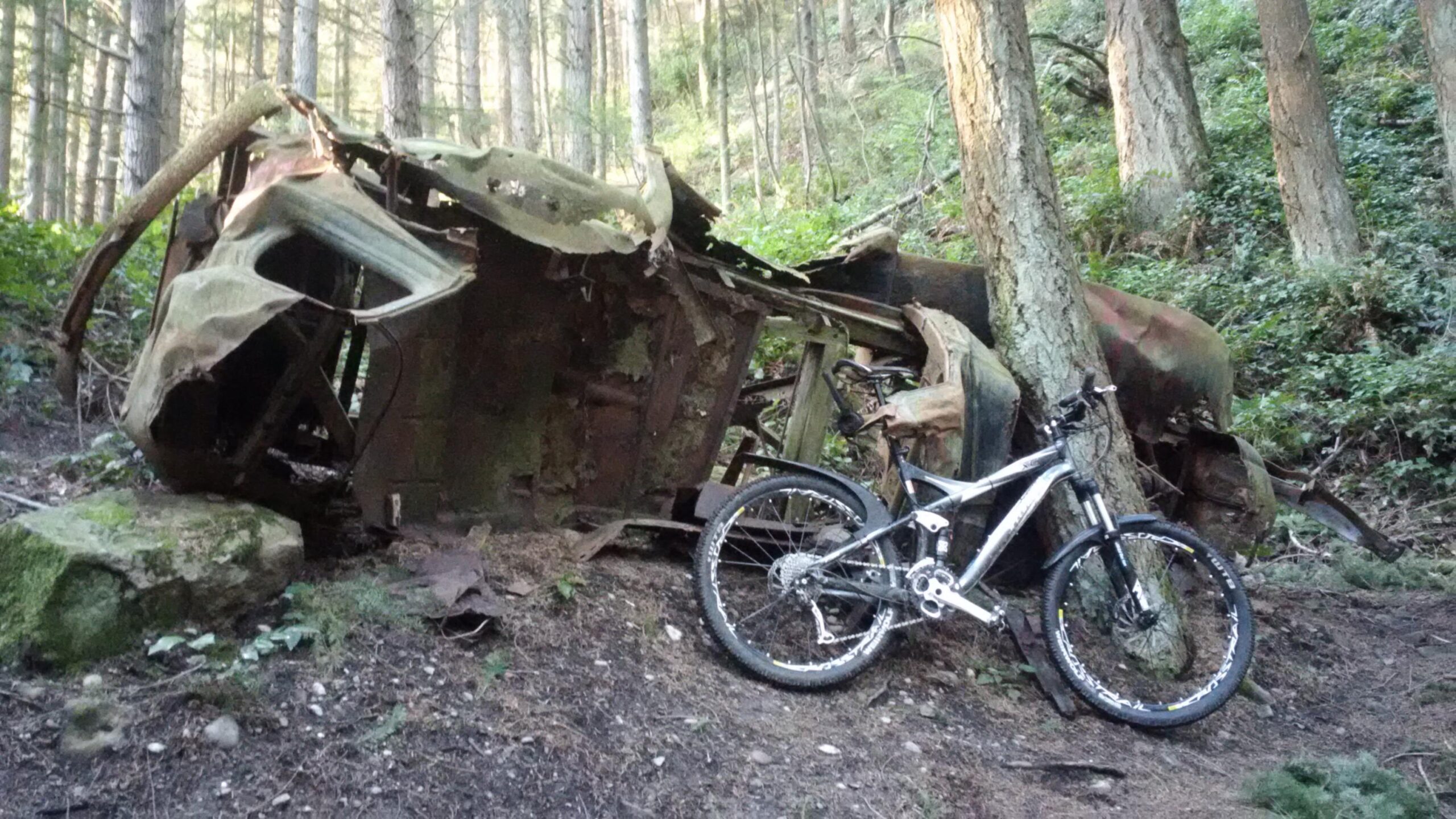 An abandoned, rusted vehicle sits partially overturned in a forested area, surrounded by trees and greenery. Next to the vehicle, a mountain bike leans against a tree, contrasting with the decaying remnants of the car. The scene captures a blend of nature and remnants of man-made structures. Little Mountain mountain bike trail.