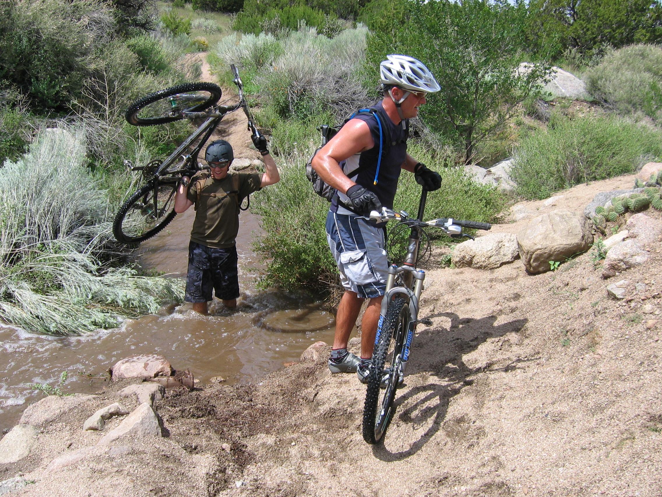 Two mountain bikers navigate a rocky trail with a stream nearby. One rider is carrying his bike over his shoulder while walking through shallow water, and the other is pedaling beside him. The landscape features dry vegetation and rocky terrain under a clear blue sky.