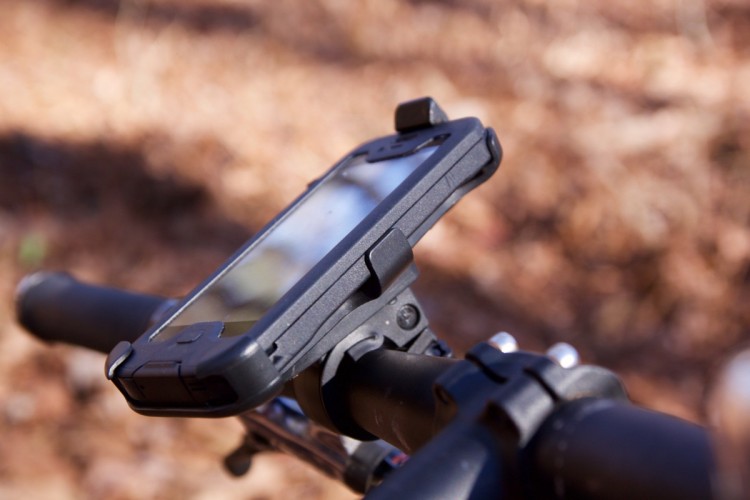A close-up image of a smartphone mounted on the handlebars of a bicycle, with a rugged phone case. The background features a blurred, natural setting with leaves.