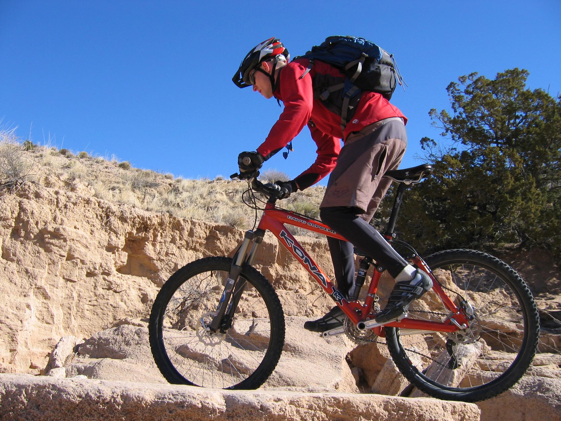 A mountain biker in a red long-sleeve shirt and black helmet navigates rocky terrain on a vibrant red bike. The background features a clear blue sky and a desert landscape with sparse vegetation.