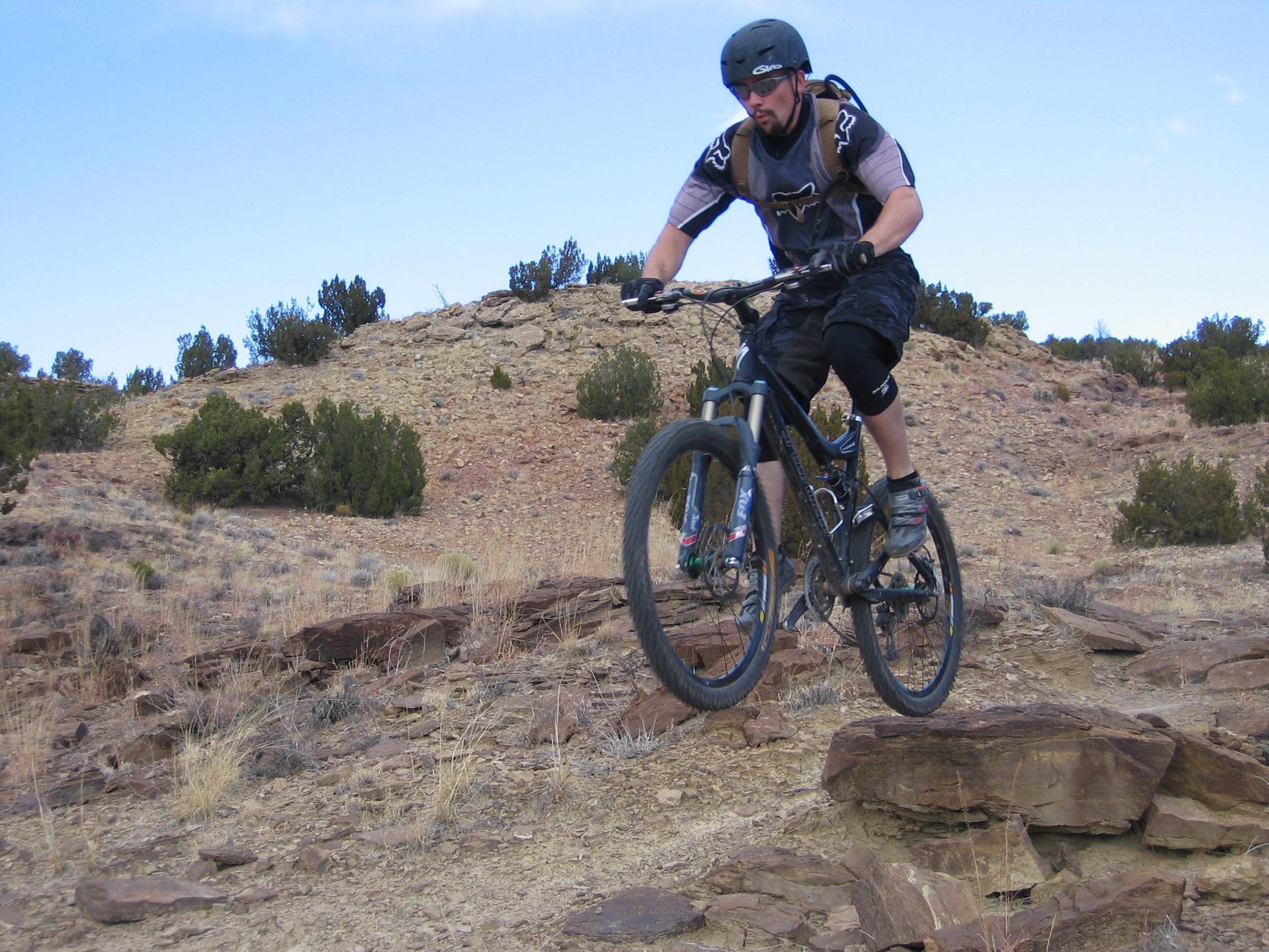 A mountain biker wearing a helmet and protective gear is performing a jump over rocky terrain, surrounded by sparse vegetation and a hilly landscape under a blue sky.
