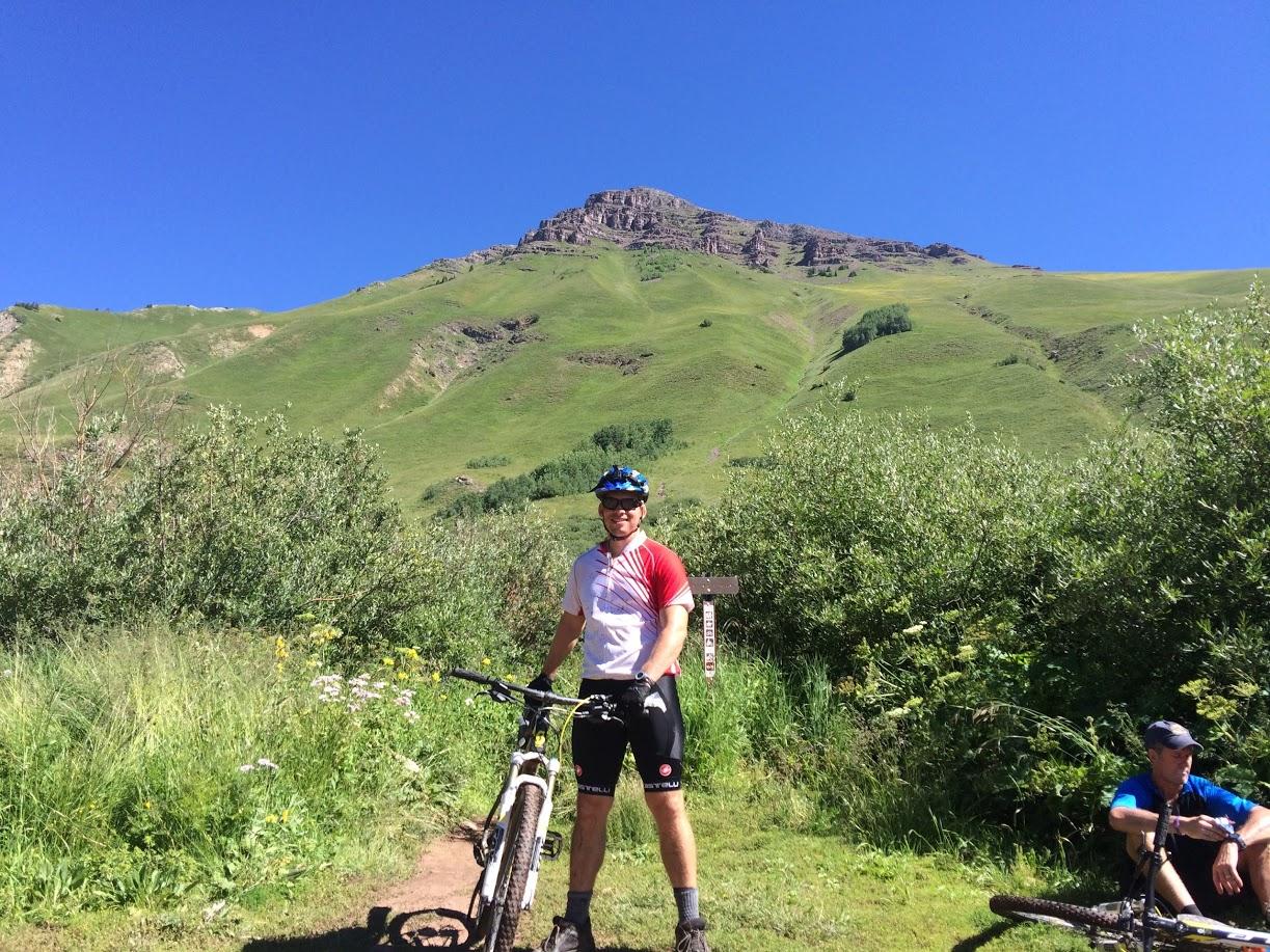 A cyclist in a red and white jersey stands with a mountain bike on a grassy trail, with a vibrant green hill and clear blue sky in the background. Another person is seated nearby, also wearing cycling gear. Teocalli Ridge mountain bike trail.