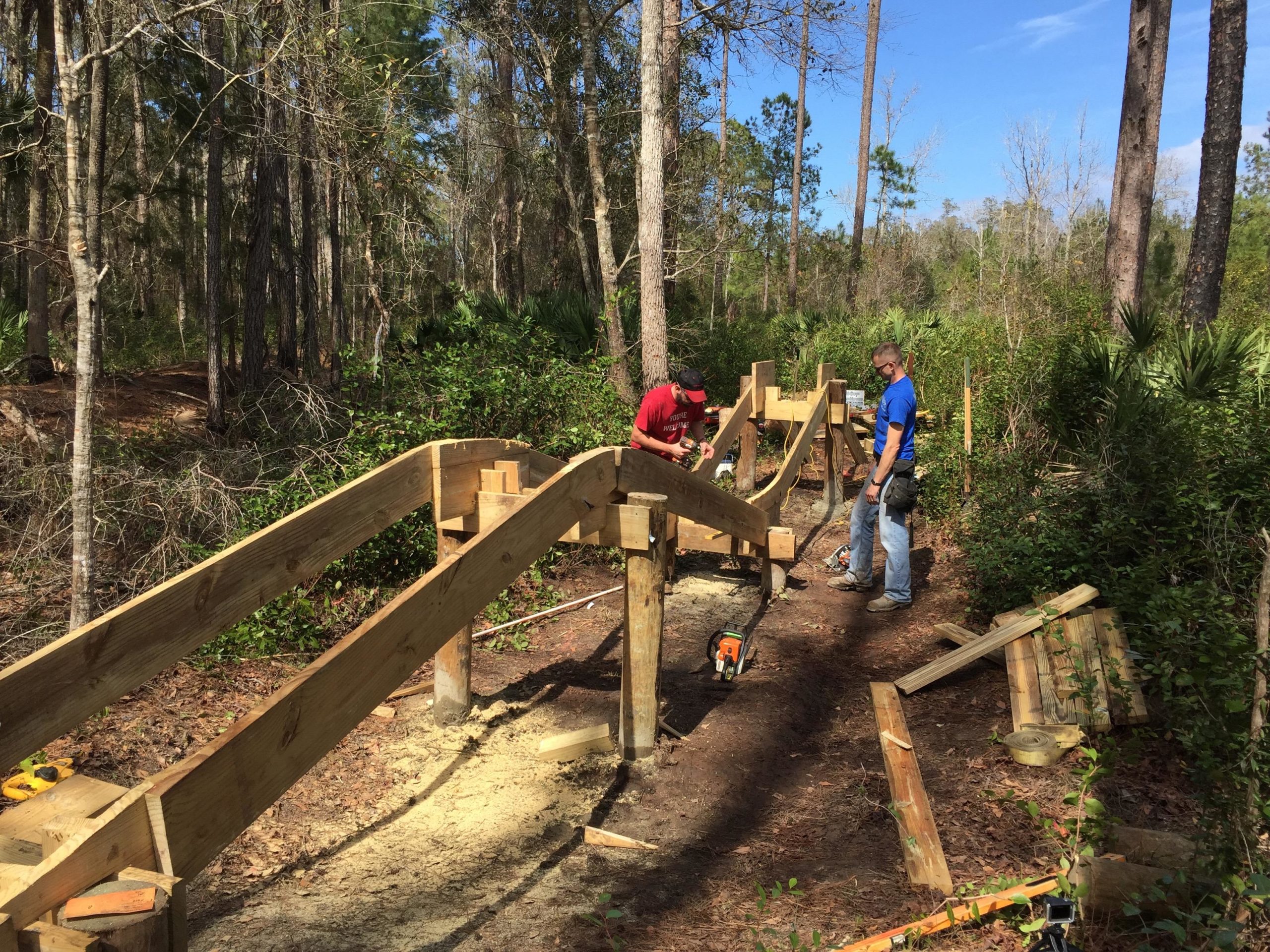 Two individuals are constructing a wooden ramp in a wooded area. One person is kneeling and examining the ramp, while the other stands nearby, holding tools. Surrounding them are trees, underbrush, and sunlight filtering through the foliage. Various tools and materials are scattered on the ground, indicating ongoing construction work. Nocatee mountain bike trail.