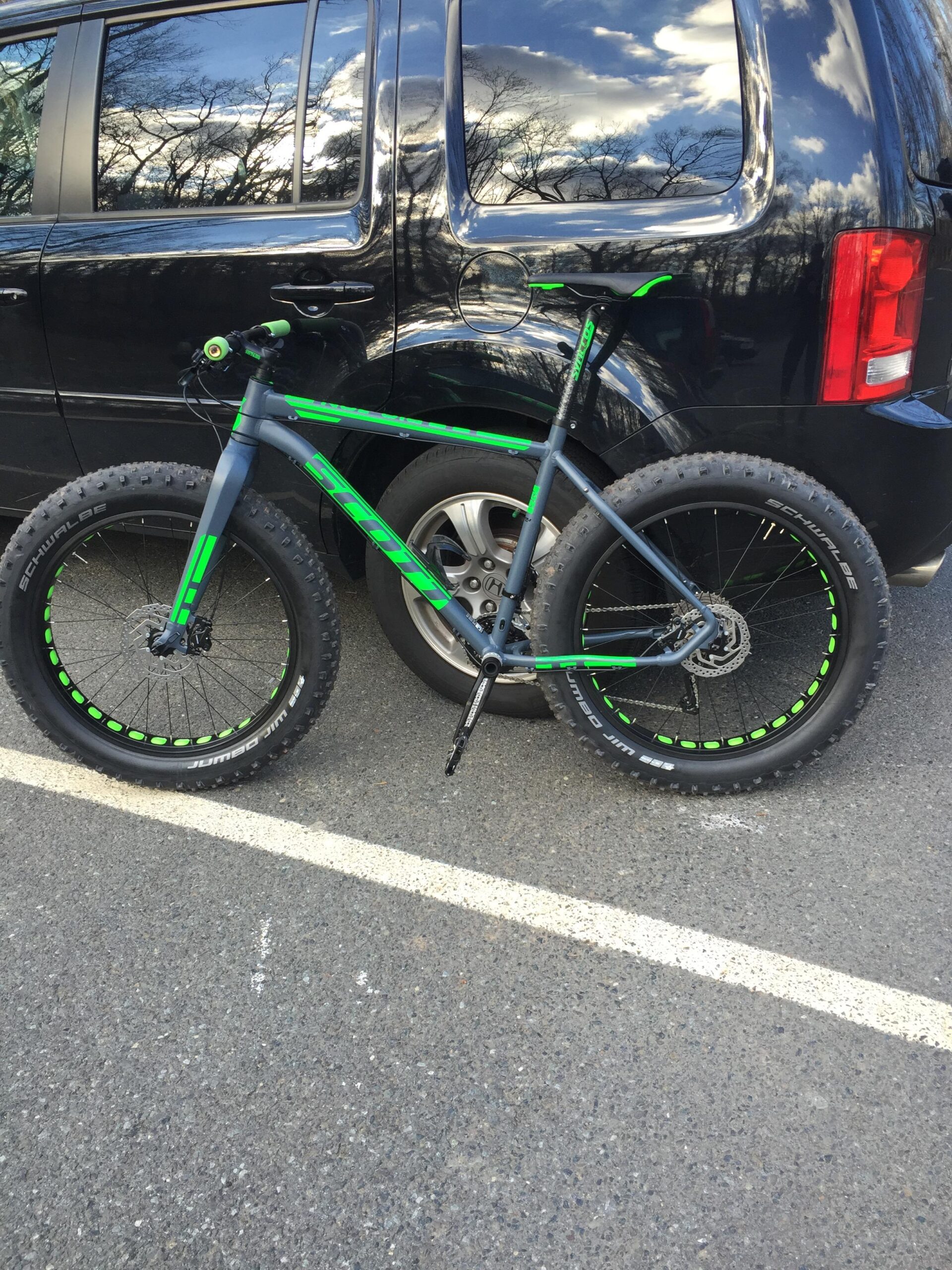 Scott Big Jon: A green and gray fat bike parked next to a black vehicle in a parking lot. The bike features wide tires and a matte finish, with green accents on the frame and wheels. The background includes a partially cloudy sky reflected in the vehicle's windows.