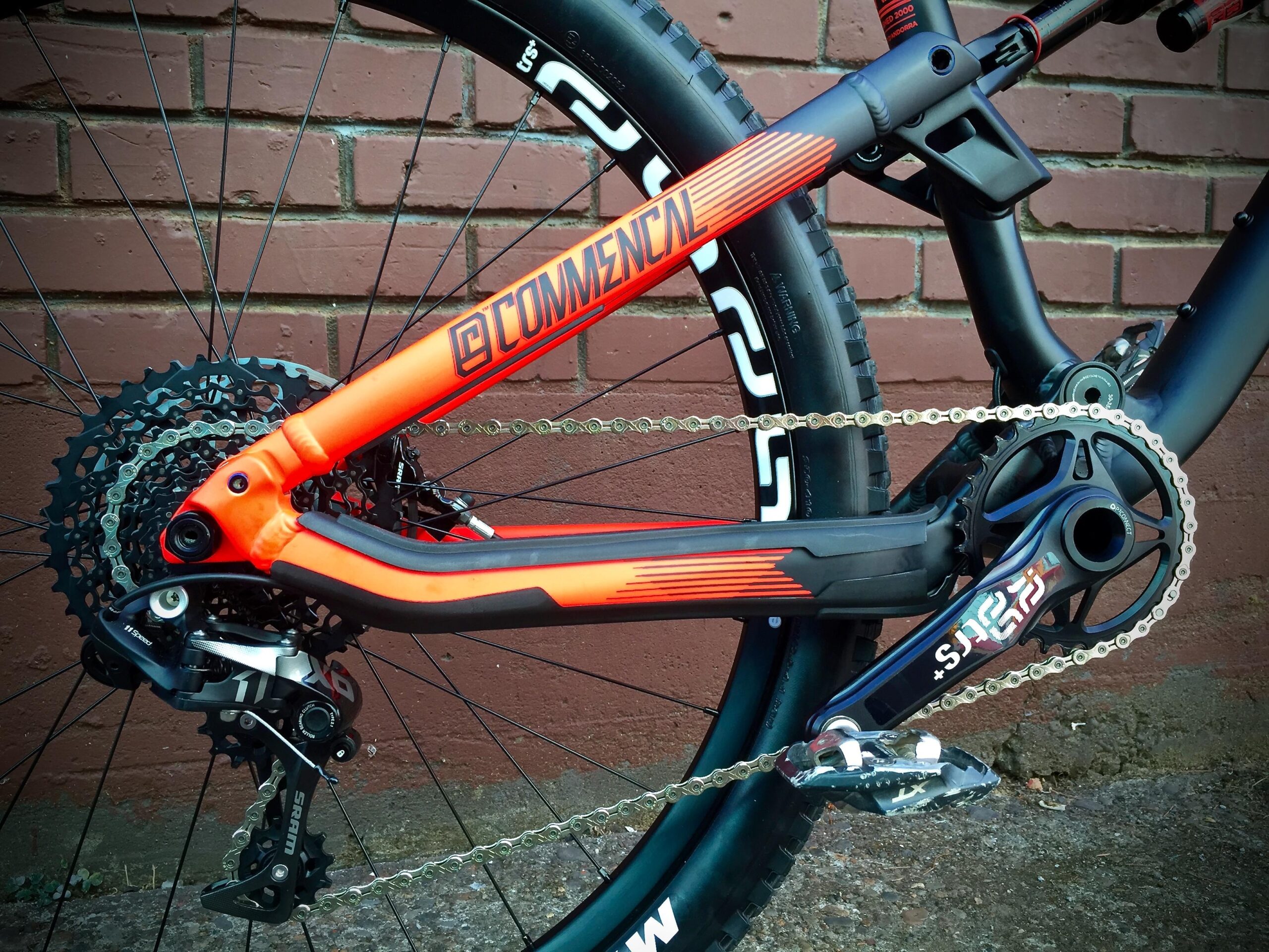 Commencal Meta AM V4: Close-up of a bicycle's drivetrain, featuring a black and orange frame, an 11-speed cassette, and a derailleur. The background consists of a brick wall, highlighting the bike's components and design.