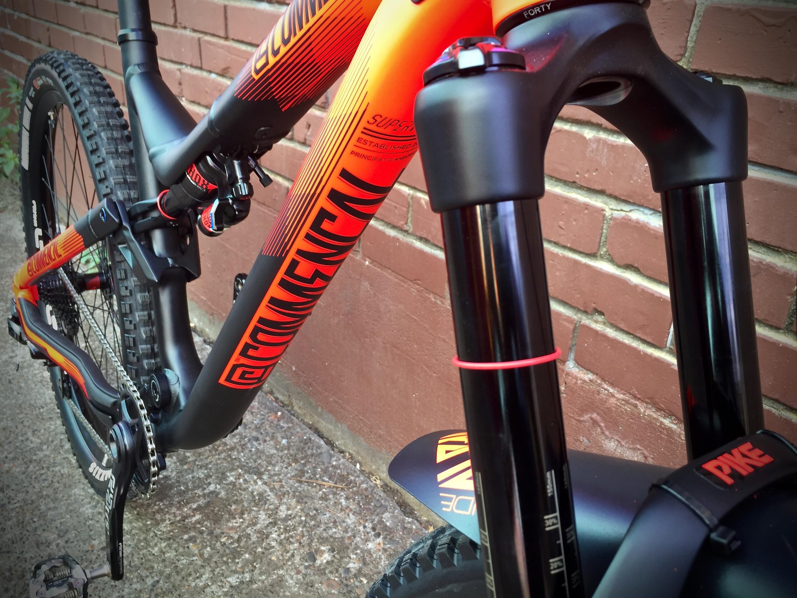 Commencal Meta AM V4: Close-up view of a mountain bike frame in vibrant orange and black, showing the handlebars, fork, and part of the drivetrain, with a brick wall in the background.