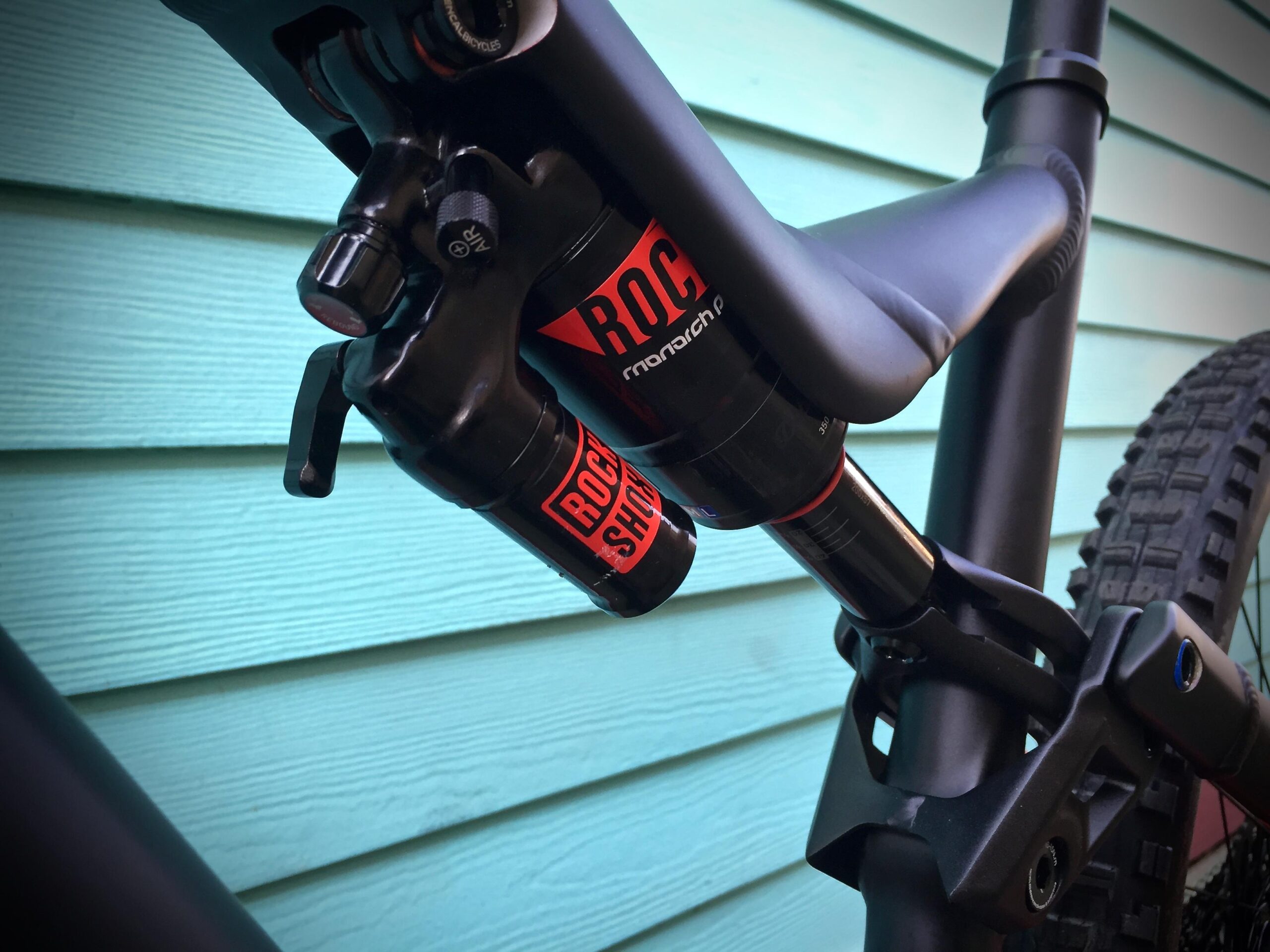 Commencal Meta AM V4: Close-up view of a bicycle's rear shock absorber, featuring a black and red RockShox design, mounted on a carbon frame. The background is a teal wall, highlighting the mechanical components of the bike.