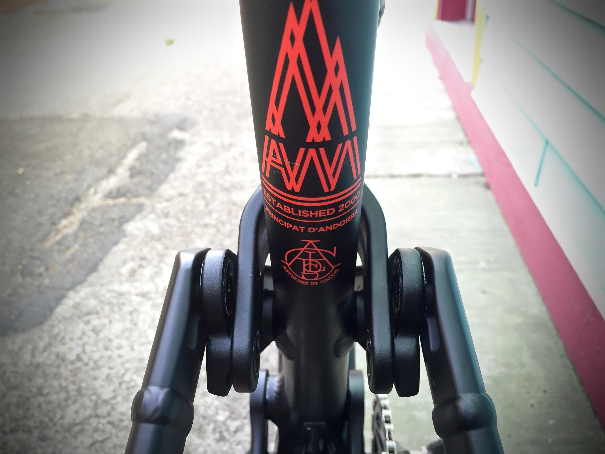 Commencal Meta AM V4: Close-up of a black bicycle frame with a distinctive red logo and text indicating the brand name and establishment year, positioned alongside a blurred outdoor pavement scene.