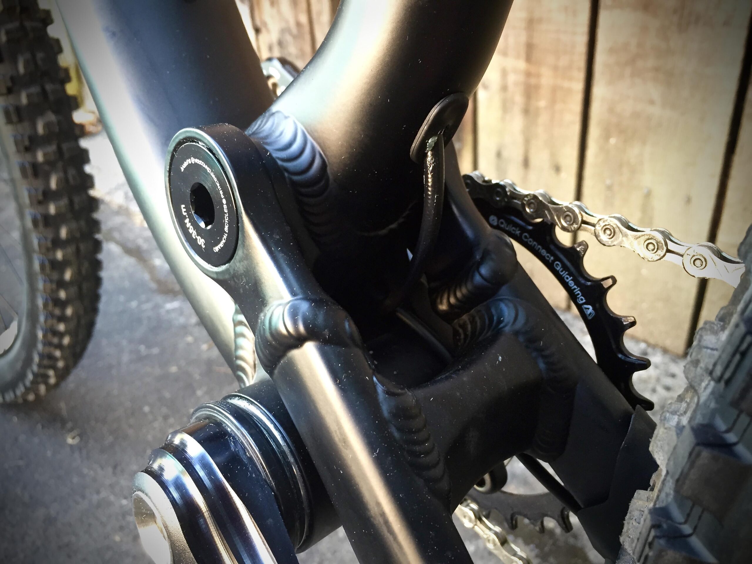 Commencal Meta AM V4: Close-up view of a mountain bike's bottom bracket area, showcasing the chainring, chain, and part of the bike frame, with a background of a wooden fence and a blurred tire.