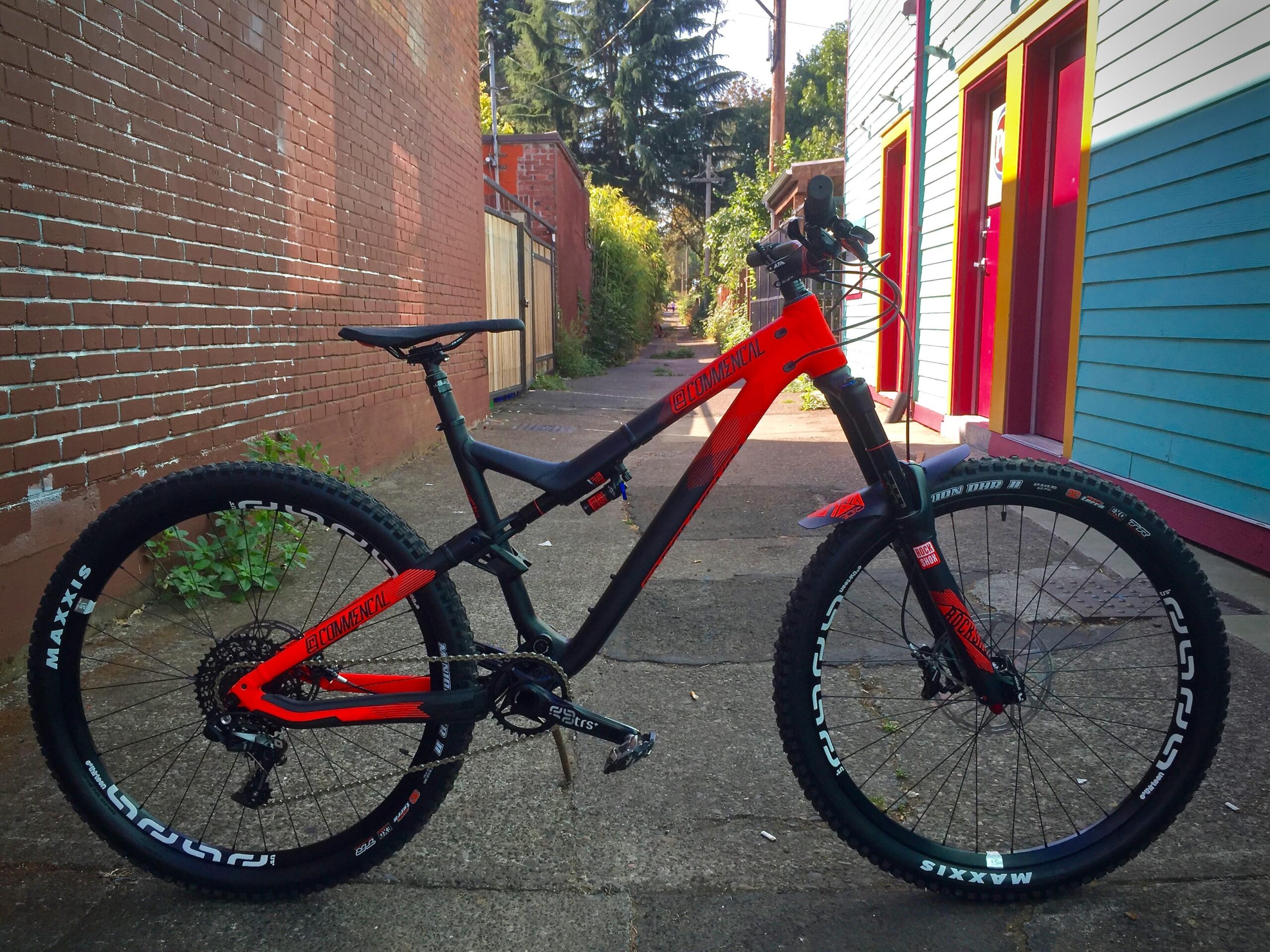 Commencal Meta AM V4: A bright orange and black mountain bike is positioned on a concrete path next to a brick wall. The bike features thick tires marked with the brand name "Maxxis" and has a sleek, modern design that highlights its suitability for off-road riding. In the background, there is a colorful building with red and blue siding, as well as greenery lining the path. The scene is set in an urban environment, partly shaded by trees.