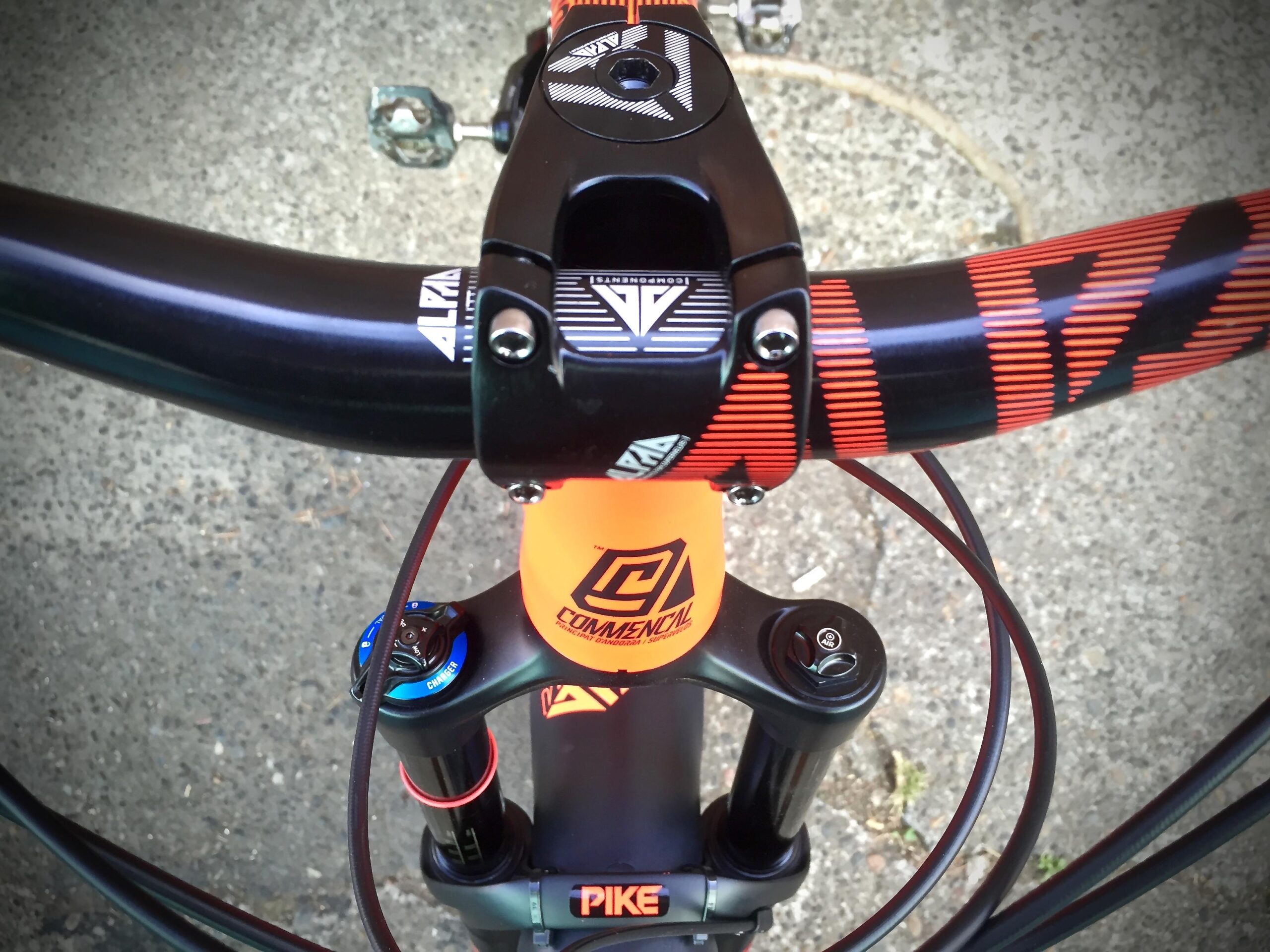 Commencal Meta AM V4: A close-up view of a mountain bike's handlebars and stem, featuring a black handlebar with red and black graphics, and an orange stem with a logo. The image also shows the bike's suspension system components and cables, set against a textured, outdoor surface.