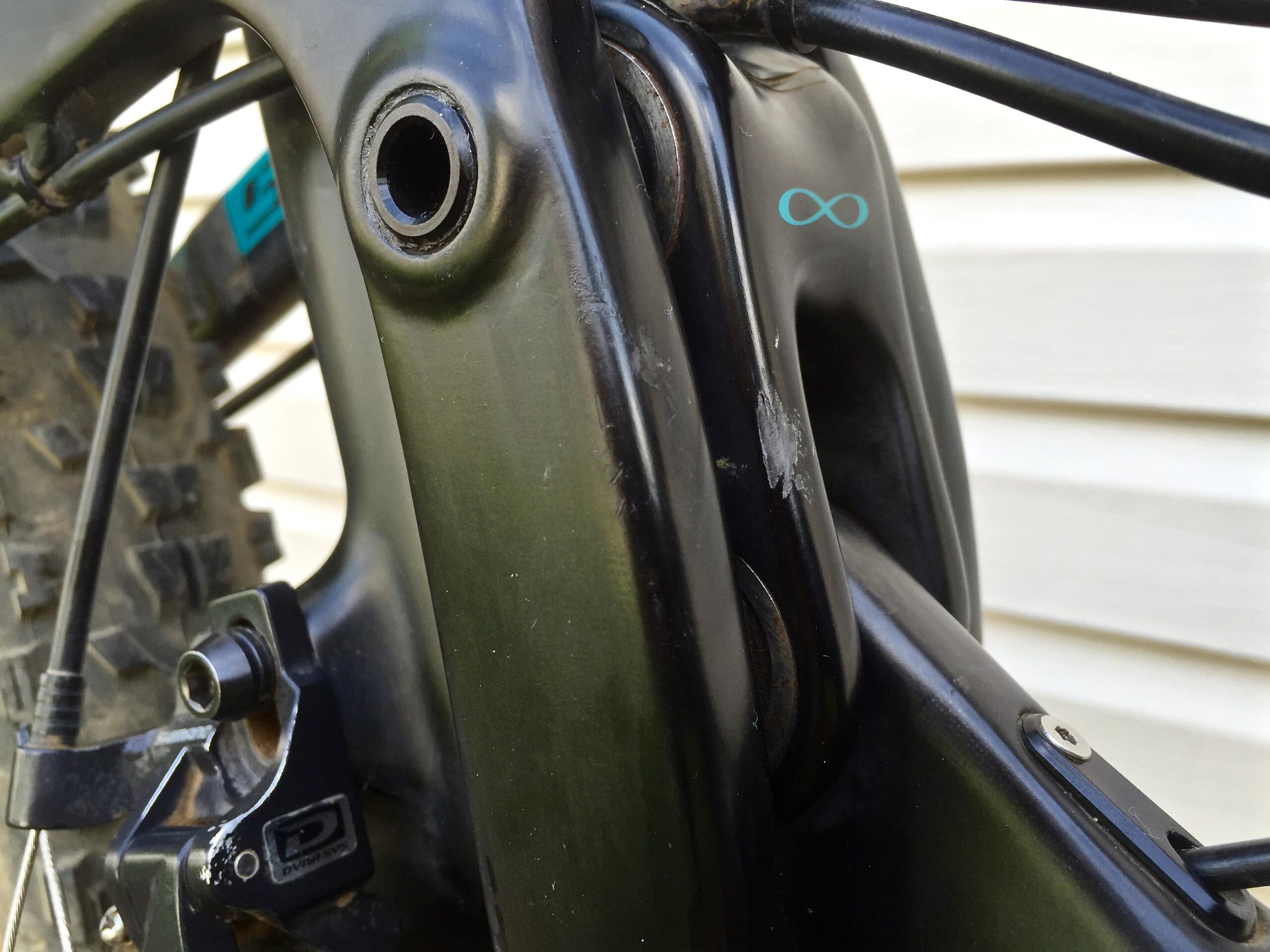 Yeti SB5c: Close-up view of a mountain bike frame, showing the area near the rear suspension linkage. The frame is a combination of black and dark green colors, with visible components such as a cable housing and a small logo featuring an infinity symbol. Some marks and scratches are apparent on the surface, indicating wear from use.