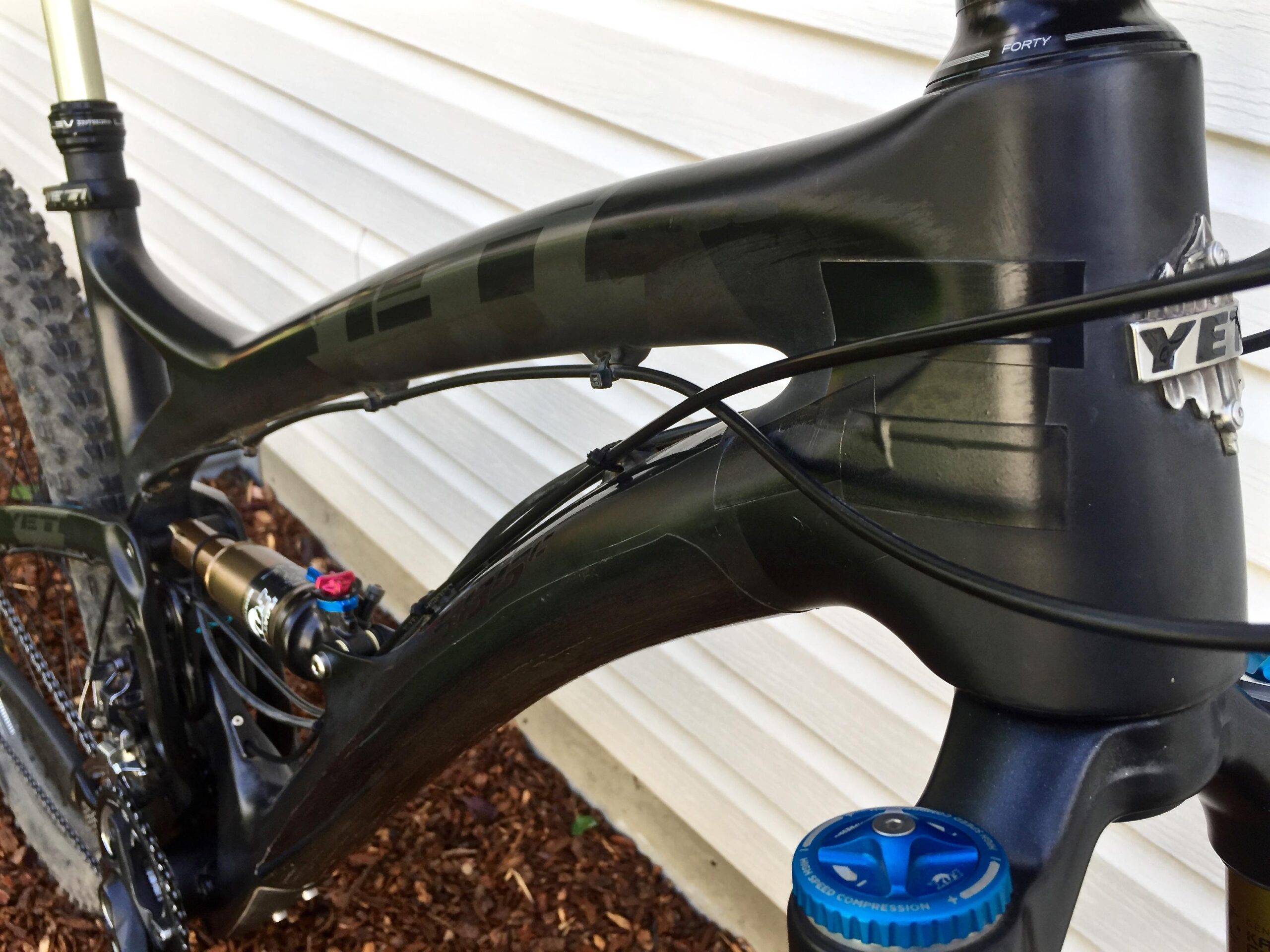 Yeti SB5c: Close-up view of a black mountain bike frame showcasing the head tube, associated cable routing, and a shock absorber with blue and pink adjustment knobs. The bike is positioned against a light-colored wall, with mulch visible in the foreground.