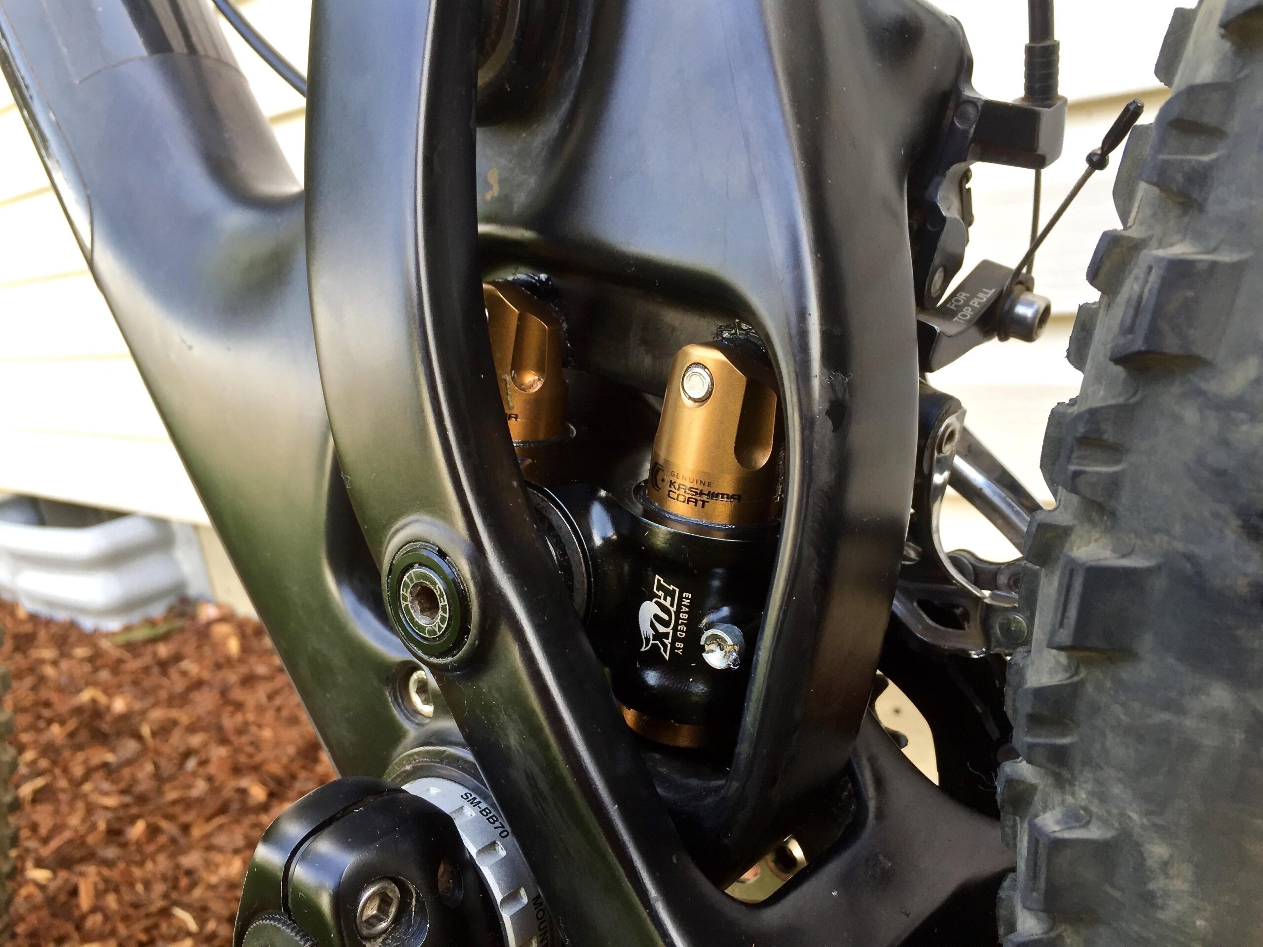 Yeti SB5c: Close-up view of a mountain bike's rear shock assembly, featuring a black frame, gold shock components, and visible adjustment knobs. The background shows a portion of a house and landscape with mulch.