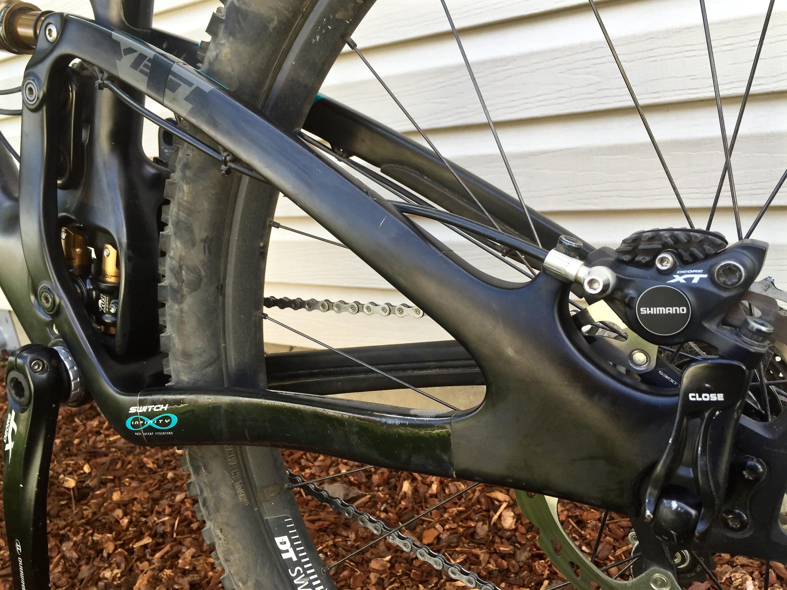 Yeti SB5c: Close-up view of a mountain bike's rear suspension and drivetrain components, featuring a Yeti bicycle frame, Shimano XT brake system, and a visible tire with knobby tread texture. The background includes wooden mulch and a textured wall.