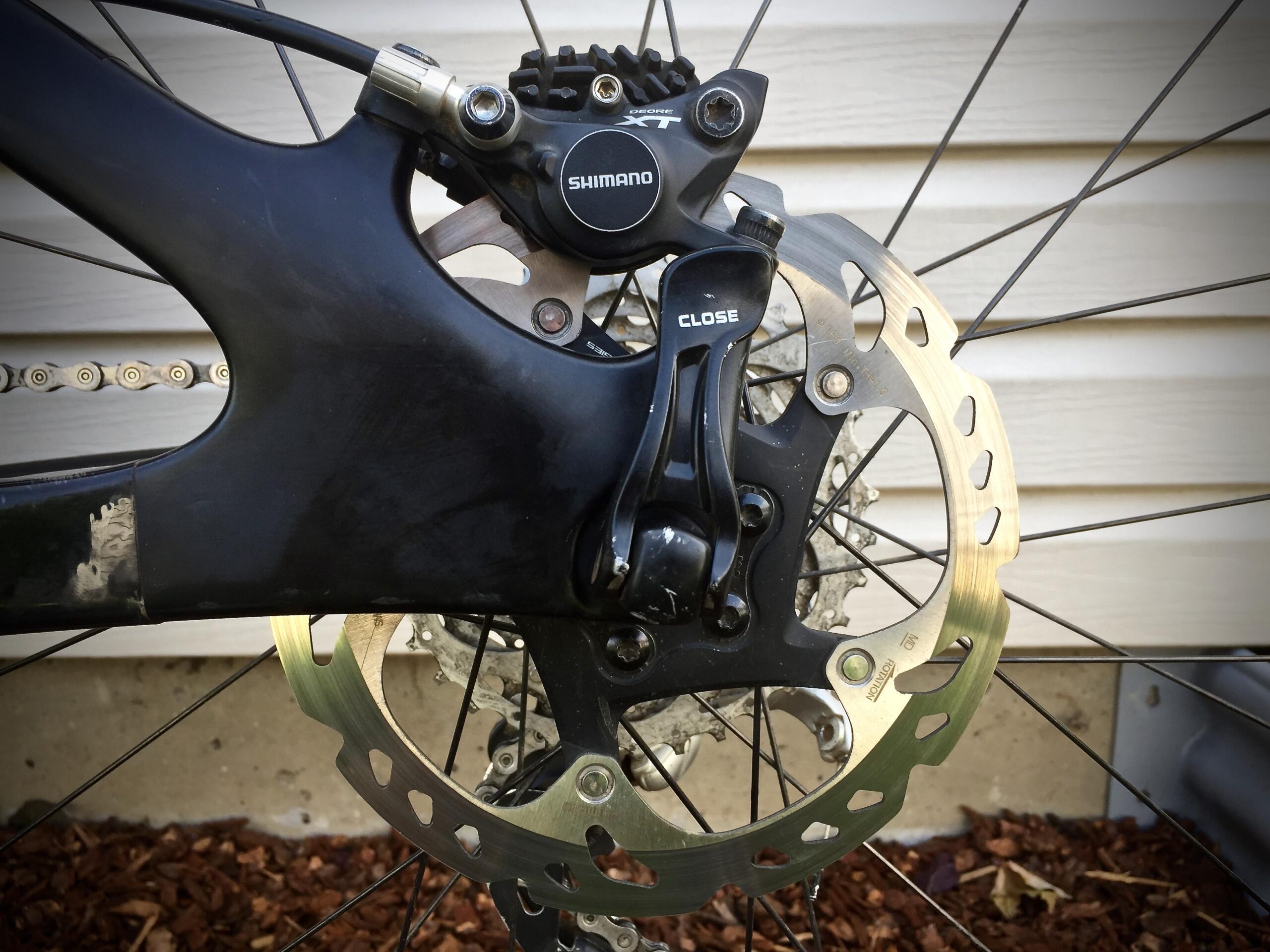 Yeti SB5c: Close-up image of a bicycle's rear disc brake system, featuring a Shimano brake caliper and rotor. The background shows parts of a bike frame and chain with wooden mulch visible below.