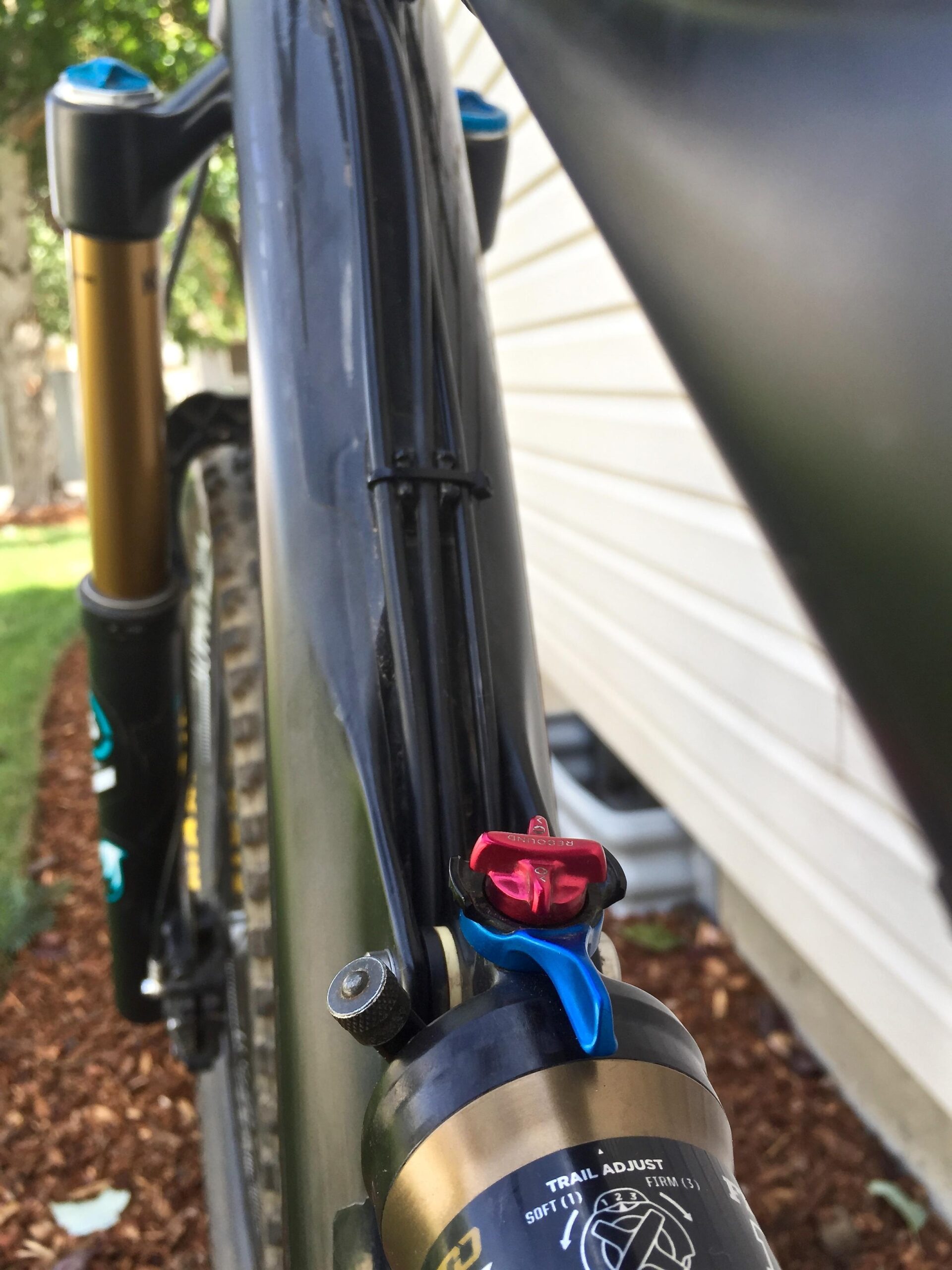 Yeti SB5c: Close-up view of a mountain bike suspension system, showcasing the shock absorber with colorful adjustment knobs and cables, set against a blurred background of outdoor greenery and a light-colored wall.