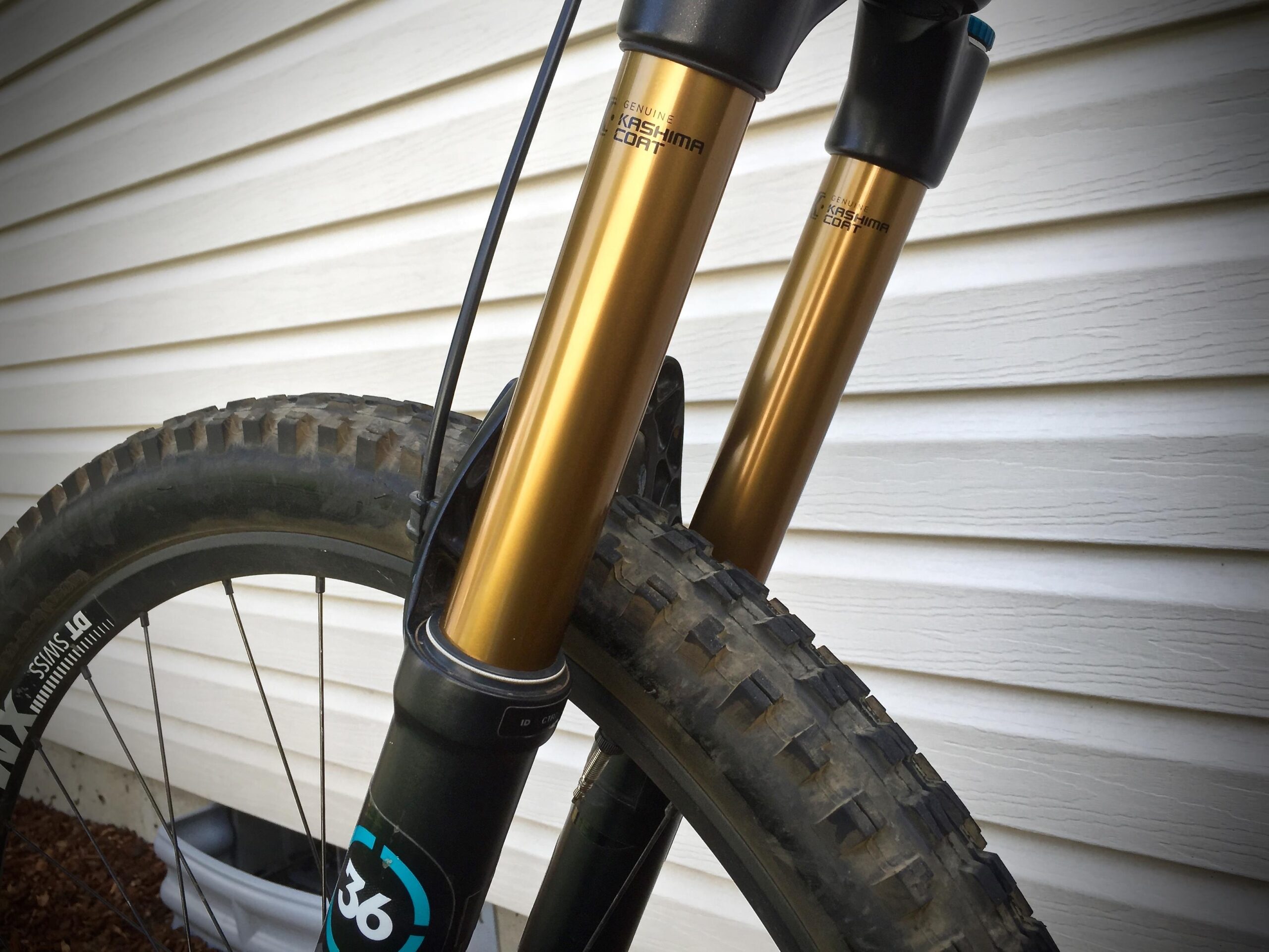 Yeti SB5c: Close-up view of a mountain bike's front suspension forks with gold anodized finish, positioned next to a textured tire. The background features a white horizontal siding.