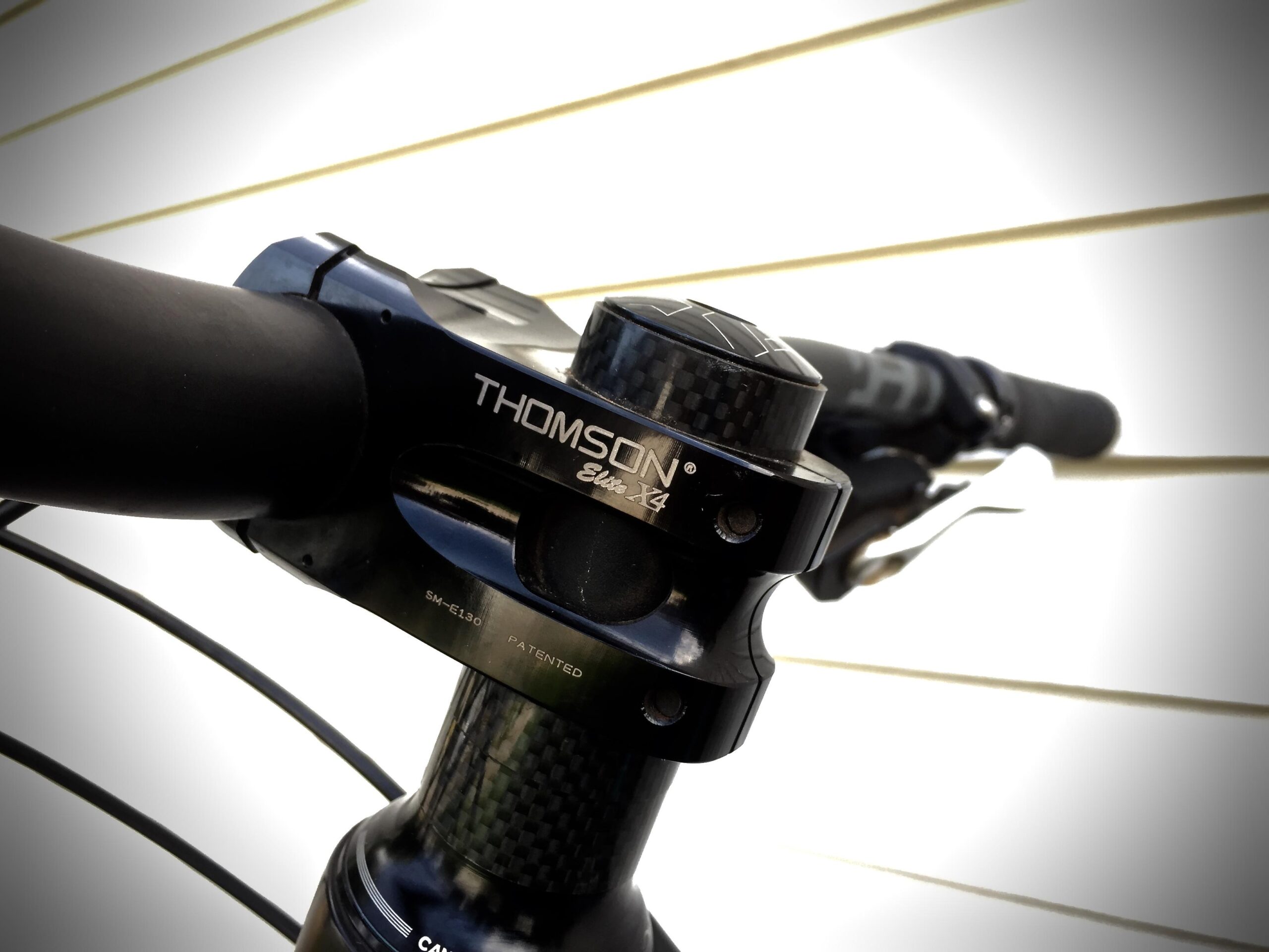 Yeti SB5c: Close-up of a Thomson Elite X4 bicycle stem attached to handlebars, featuring a sleek black finish and carbon fiber details, with a blurred background of vertical lines indicating a wall or surface.