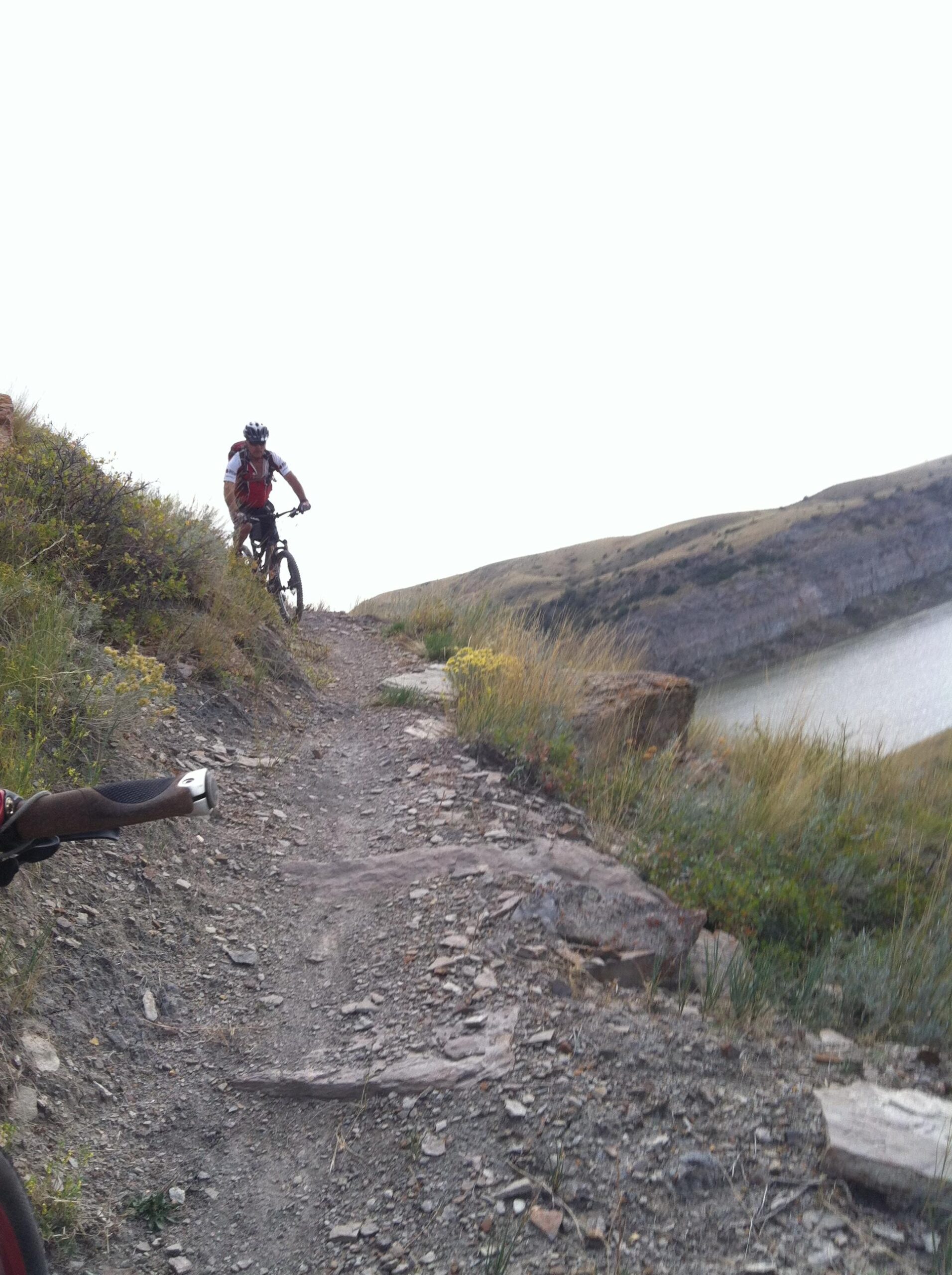 A mountain biker navigating a rocky, narrow trail along a steep hillside with grassy vegetation and a view of a lake below. The background features rolling hills under a cloudy sky. New Video Game/Yeah Kid mountain bike trail.