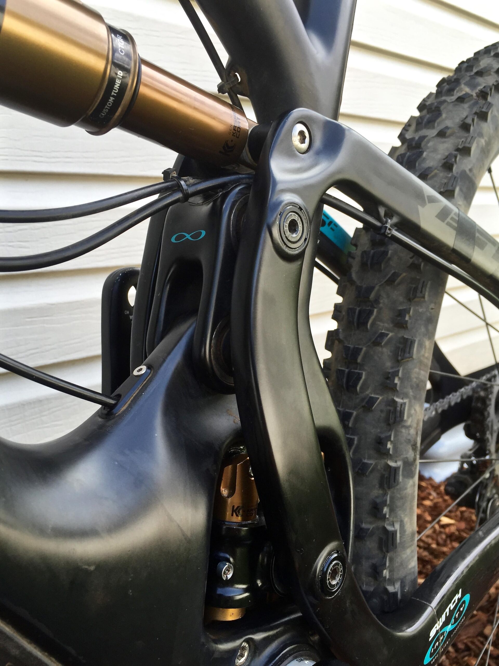 Yeti SB5c: Close-up view of a bicycle's rear shock and suspension linkage. The image shows a black frame with gold and green shock components, highlighting the intricate design and engineering of the suspension system. Thin cables run alongside the frame, and the background features a textured white wall.