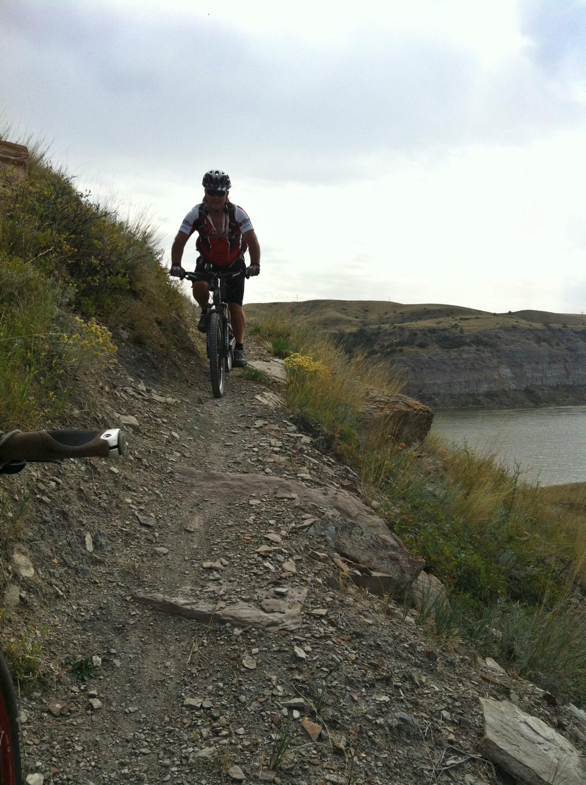 A mountain biker rides along a narrow, rocky trail above a body of water, surrounded by grassy hills and a cloudy sky. The biker is wearing a helmet and a short-sleeved jersey, focused on navigating the challenging terrain. New Video Game/Yeah Kid mountain bike trail.