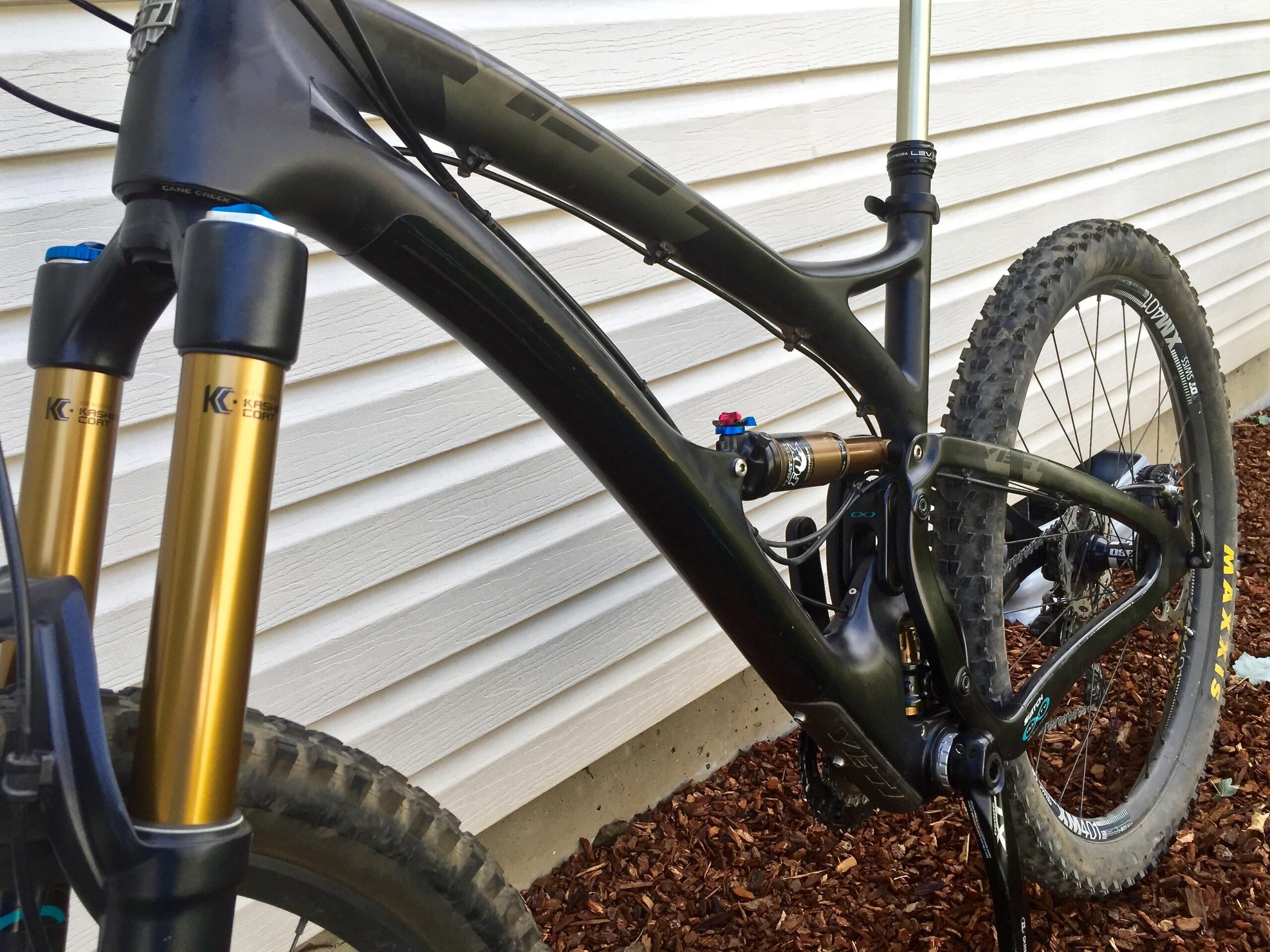Yeti SB5c: A close-up view of a modern mountain bike showcasing its black frame and gold suspension forks. The background features white exterior siding and decorative mulch. The bike is equipped with high-performance components, indicating suitability for rugged terrains.