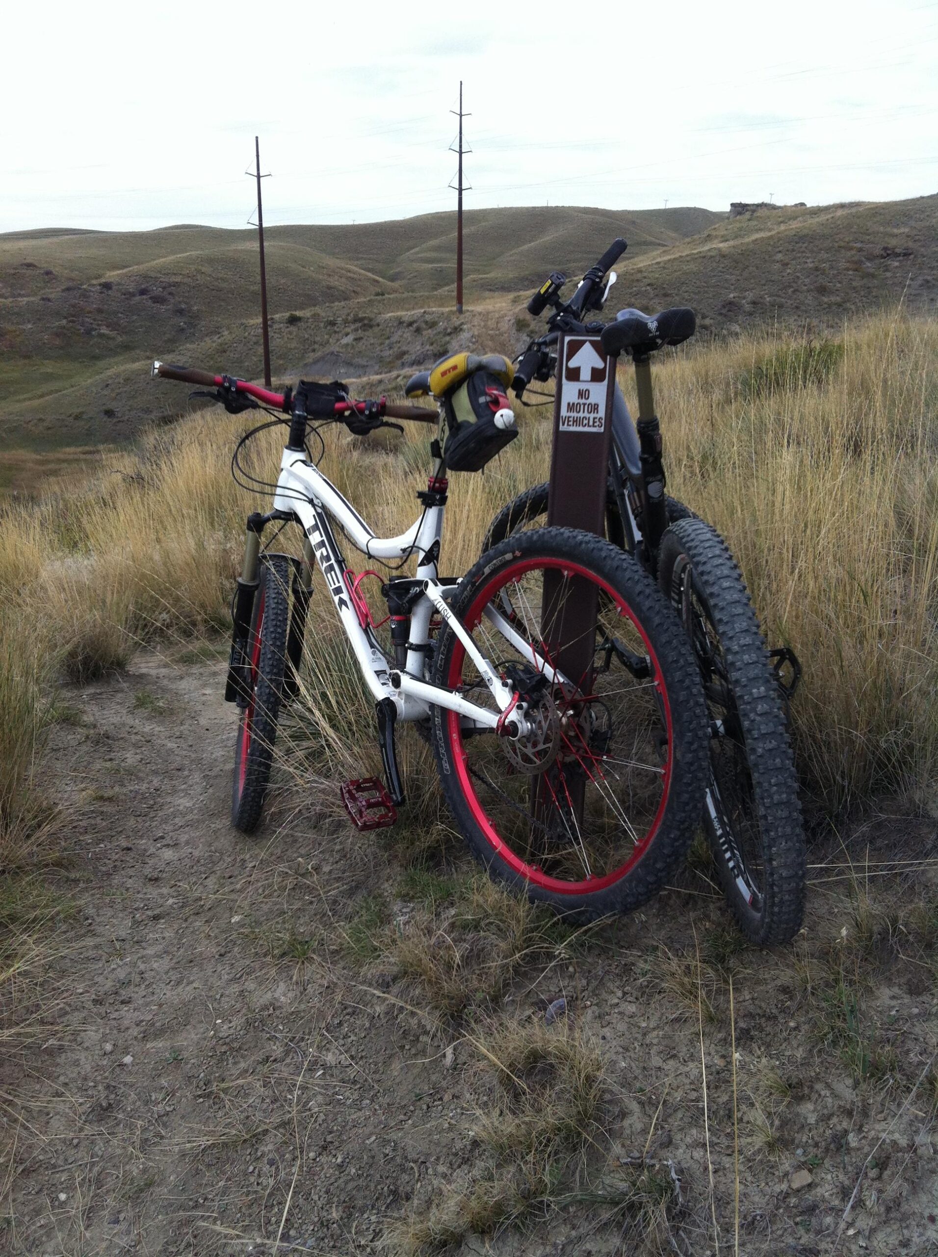 Two mountain bikes parked next to a trail sign indicating "No Motor Vehicles." The bikes, one with a white frame and red accents and the other in dark colors, rest on a dirt path surrounded by tall grass and rolling hills in the background under an overcast sky. New Video Game/Yeah Kid mountain bike trail.