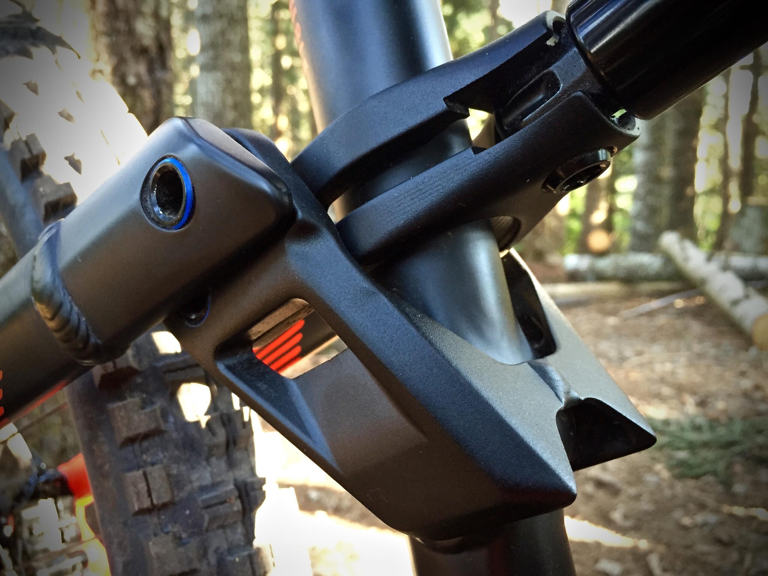 Commencal Meta AM V4: Close-up of a mountain bike frame component, focusing on the junction of the seat post and frame with a black and blue design. The background features blurred trees and natural terrain, suggesting an outdoor setting.
