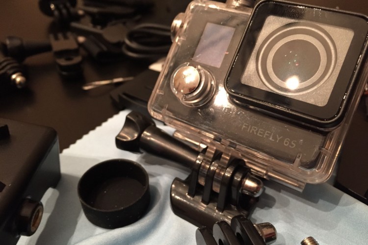 A close-up view of a Firefly 6S action camera resting on a table. Surrounding the camera are various accessories, including mounts and cables, on a blue microfiber cloth.