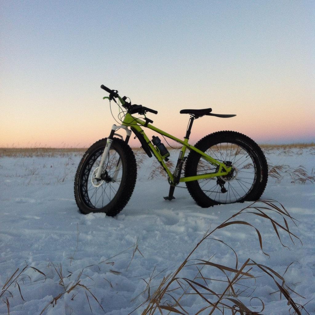 Specialized Fatboy Pro: A fat tire bike parked on snow-covered ground, with a soft gradient sky in the background transitioning from pale blue to pink, indicating dawn or dusk. Some dried grass is visible in the foreground.