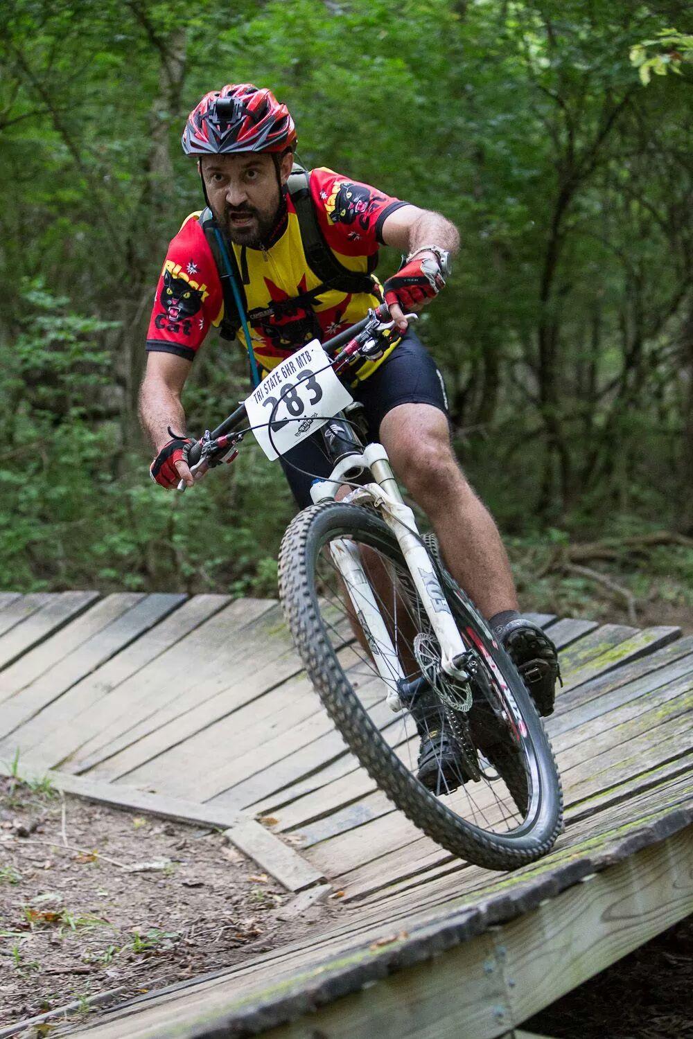A mountain biker navigating a wooden ramp on a forest trail, wearing a helmet and a brightly colored jersey, with a race number displayed on the front of the bike. The scene is surrounded by lush green foliage. England Idlewild Mountain Biking Park mountain bike trail.