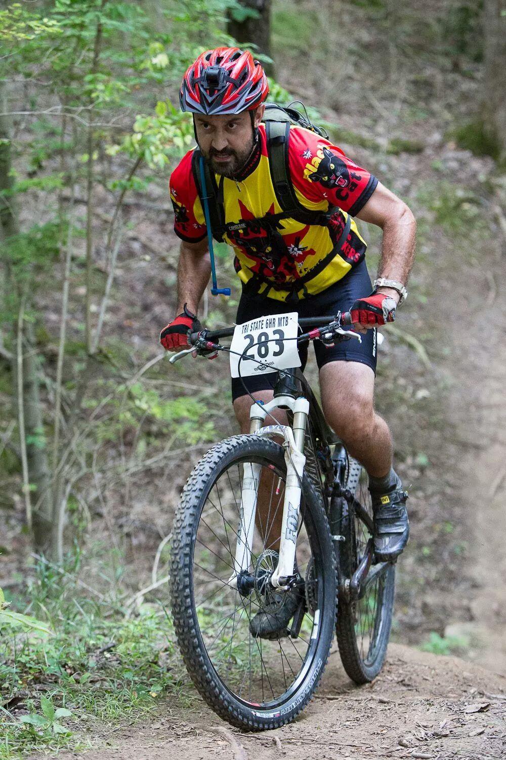 A male mountain biker wearing a colorful jersey and helmet climbs a dirt trail surrounded by trees. He is focused and determined as he rides a bicycle with a visible race number (283) attached to the front. The terrain is rugged, showcasing a natural outdoor setting. England Idlewild Mountain Biking Park mountain bike trail.