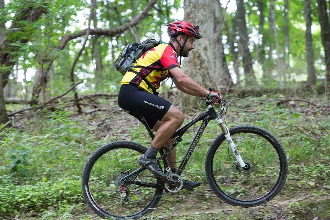A mountain biker in a colorful jersey rides a bike uphill through a forested trail, surrounded by greenery and trees. The cyclist is wearing a helmet and a backpack, demonstrating an active outdoor lifestyle. England Idlewild Mountain Biking Park mountain bike trail.
