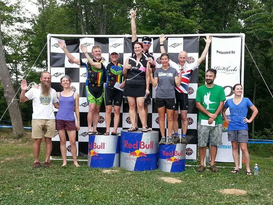 A group of eight individuals stands on a podium celebrating a cycling event. The top three competitors, dressed in cycling gear, are on raised sections of the podium holding trophies and cheering. The second and third-place finishers are wearing colorful jerseys, while the winners have their arms raised in victory. Others in the group, dressed casually, are smiling and applauding from the ground. The backdrop features a branded banner with logos and a black and white checkered pattern. The scene takes place outdoors, surrounded by greenery. England Idlewild Mountain Biking Park mountain bike trail.