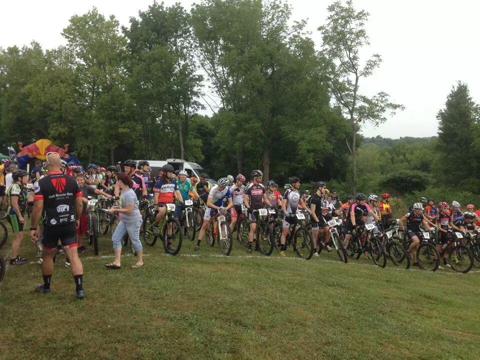 A large group of mountain bikers in colorful jerseys and helmets are lined up on a grassy field, preparing for a race. Spectators are present, and trees are visible in the background, creating a natural setting. The atmosphere is lively with participants gathered around their bicycles. England Idlewild Mountain Biking Park mountain bike trail.