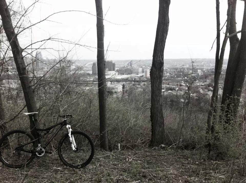 A mountain bike resting on the ground among sparse trees, with a misty cityscape in the background. The scene captures a blend of nature and urban landscape, featuring a cloudy sky and buildings in the distance. Devou Park mountain bike trail.