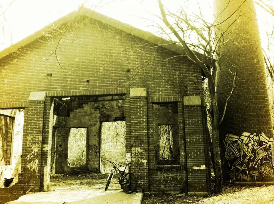 Alt text: A rustic, abandoned brick building with large openings, surrounded by overgrown trees. A bicycle is propped against the wall. A tall, cylindrical structure is visible on the right, and graffiti covers parts of the building's exterior. The image has a muted yellow tint. Devou Park mountain bike trail.
